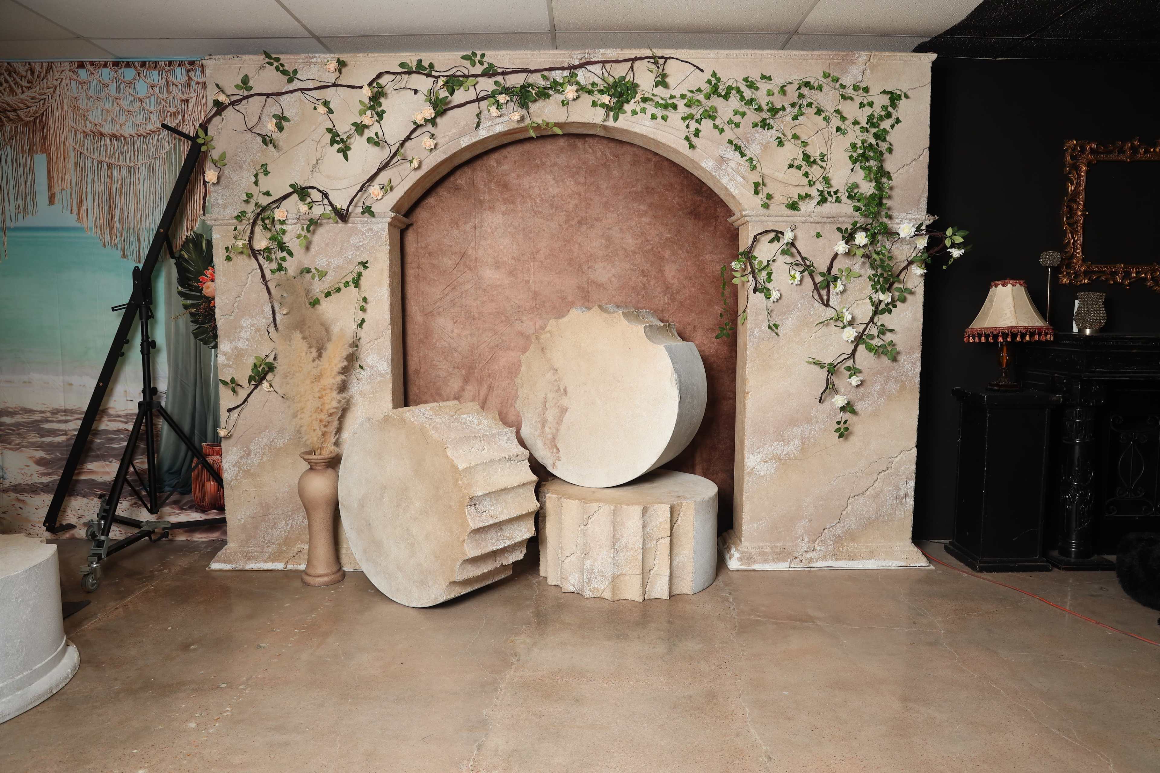 The image shows a decorative backdrop featuring an archway with a textured surface, flanked by plants, and three cylindrical pedestals in varying heights placed on the floor.
