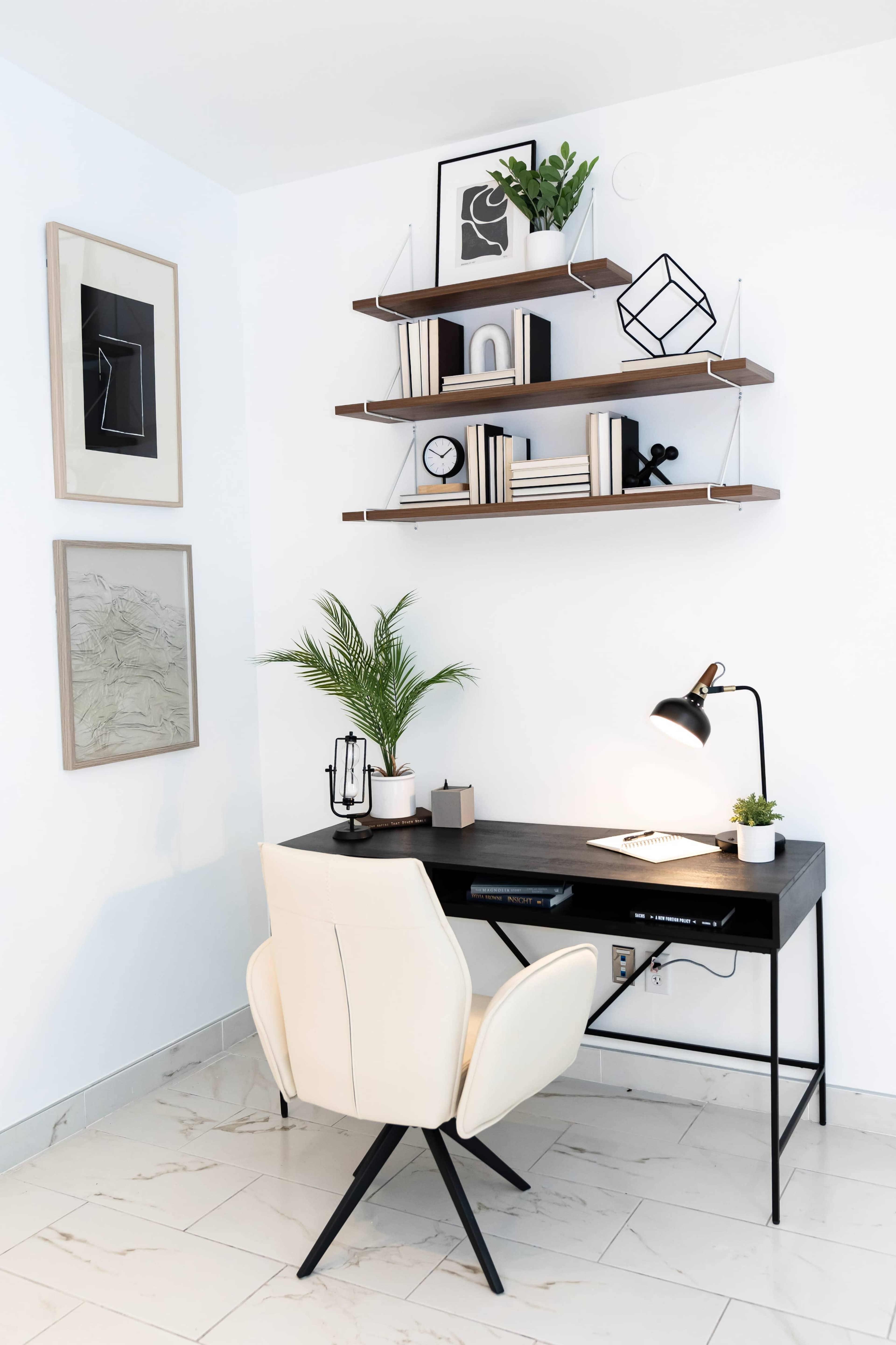 The image shows a minimalist workspace featuring a black desk, a cream-colored chair, and decorative shelves with plants and artwork on a white wall.