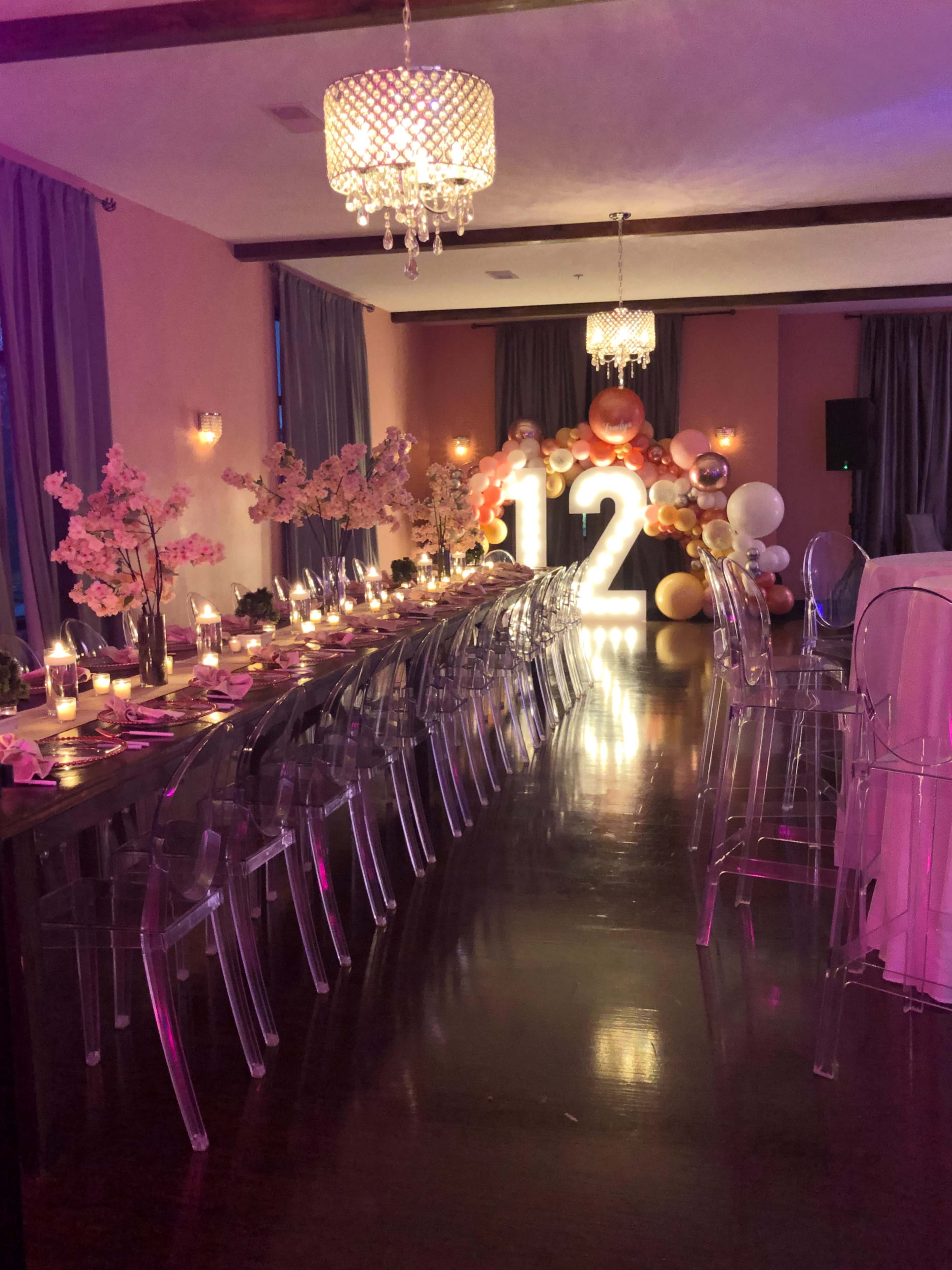 A decorated banquet hall featuring a long dining table with floral arrangements, illuminated number lights reading "12," and a backdrop of balloons.