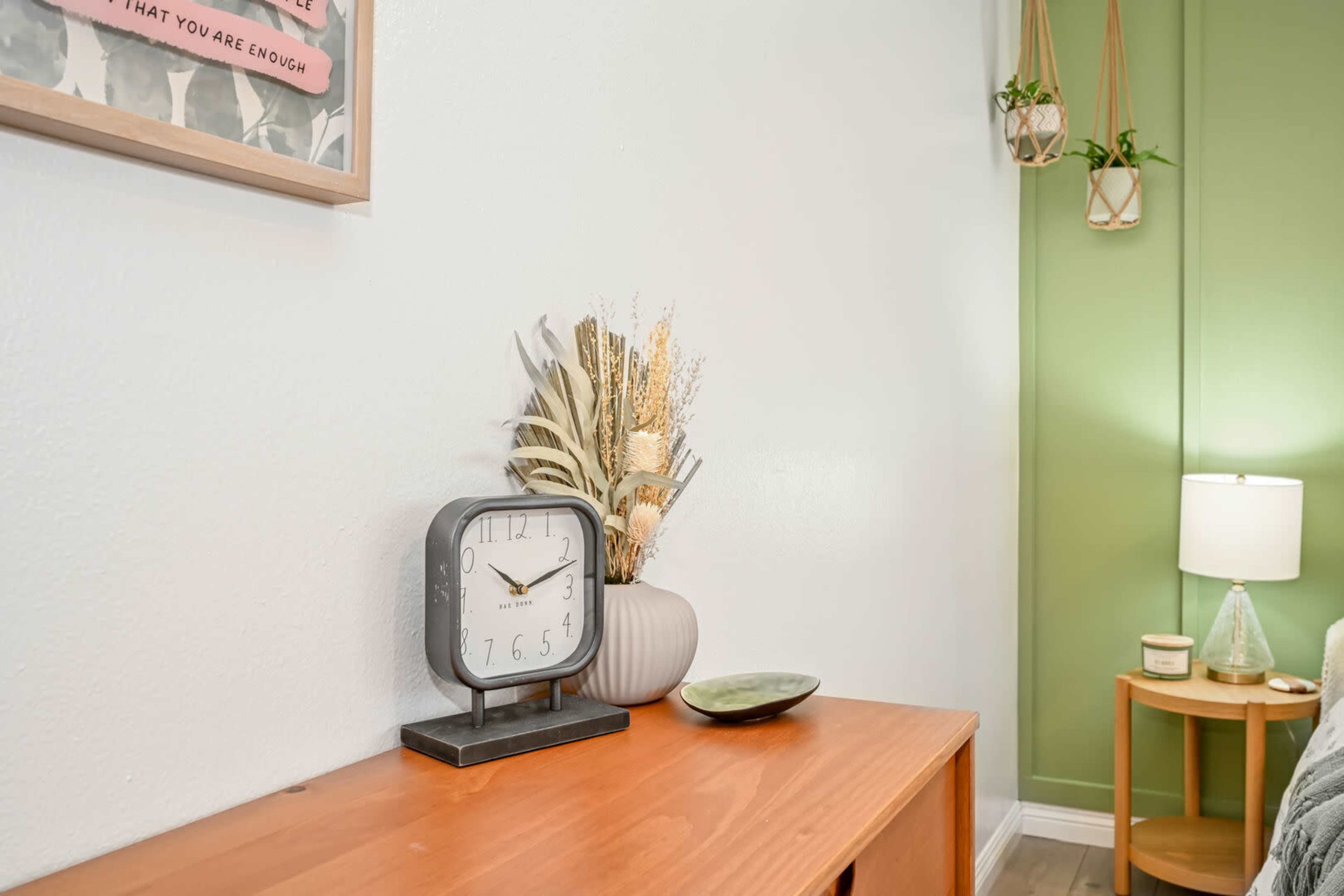 A wooden side table displays a clock, a decorative vase with dried plants, and a bowl, against a wall featuring a green accent section and a framed poster.