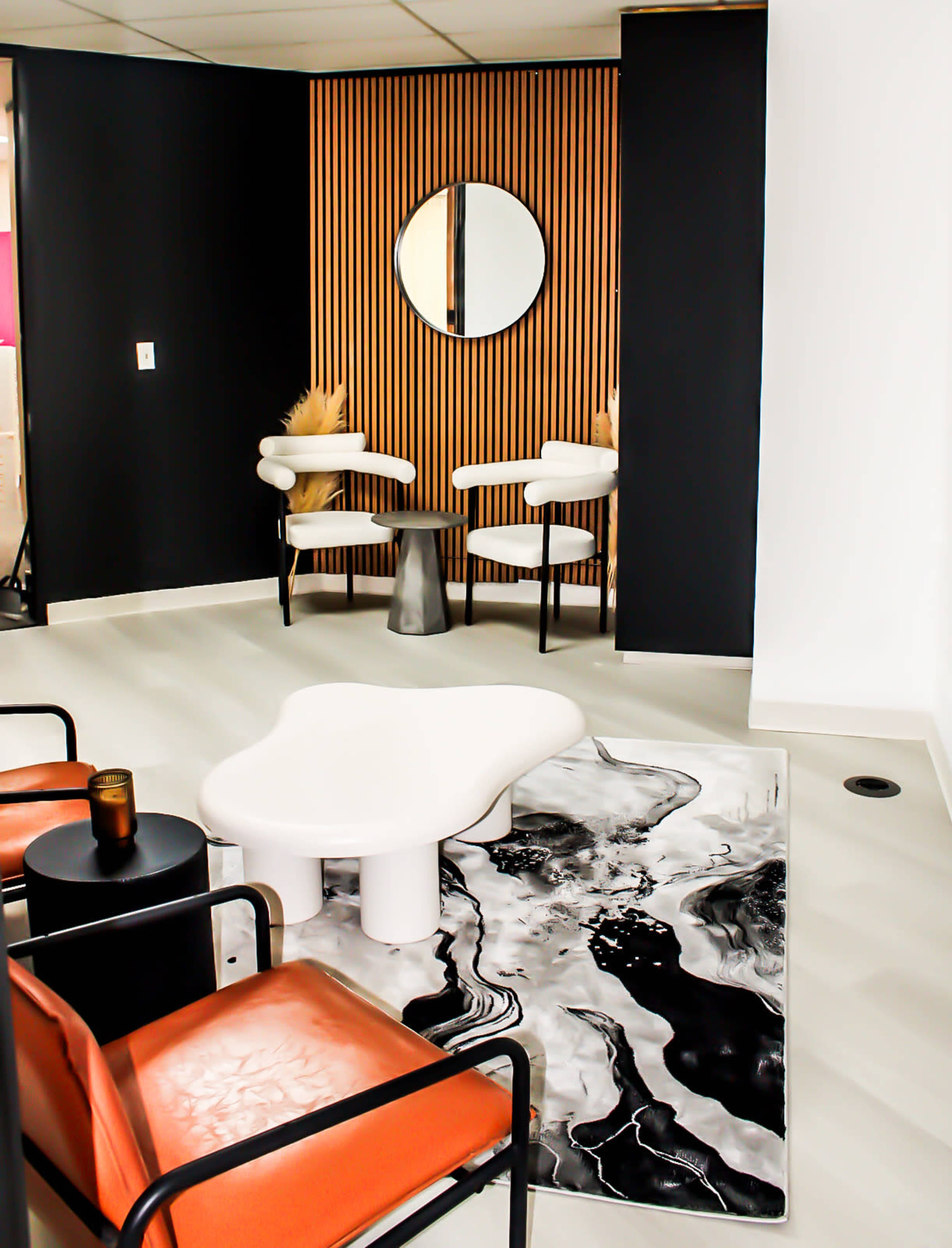 The image depicts a modern lounge area with two white armchairs and a unique coffee table on a patterned rug, complemented by a circular mirror and a minimalist decor style.