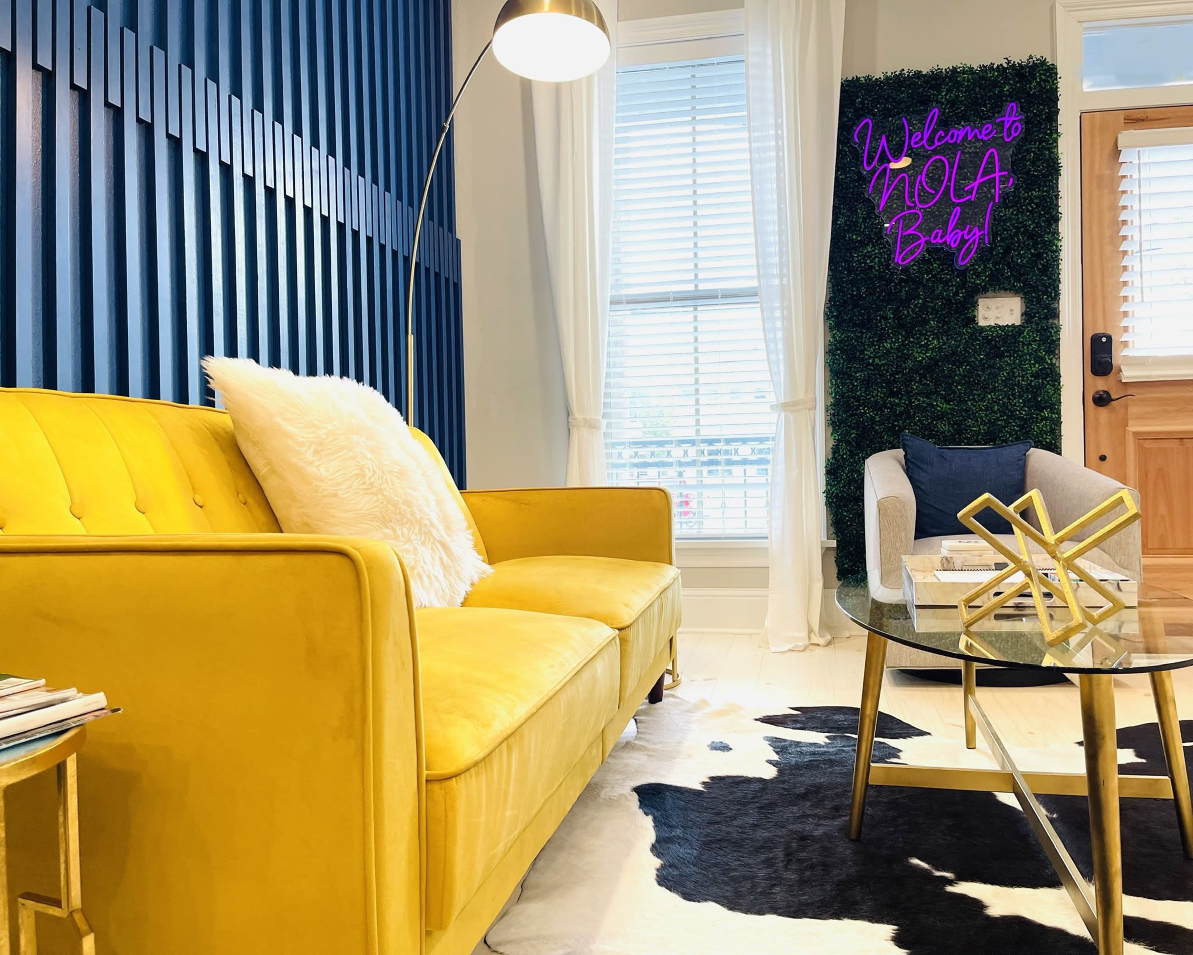 A bright yellow sofa sits in a modern room with blue striped walls, a glass coffee table on a cowhide rug, and a neon sign reading "Welcome to NOLA Baby!" against a green backdrop.