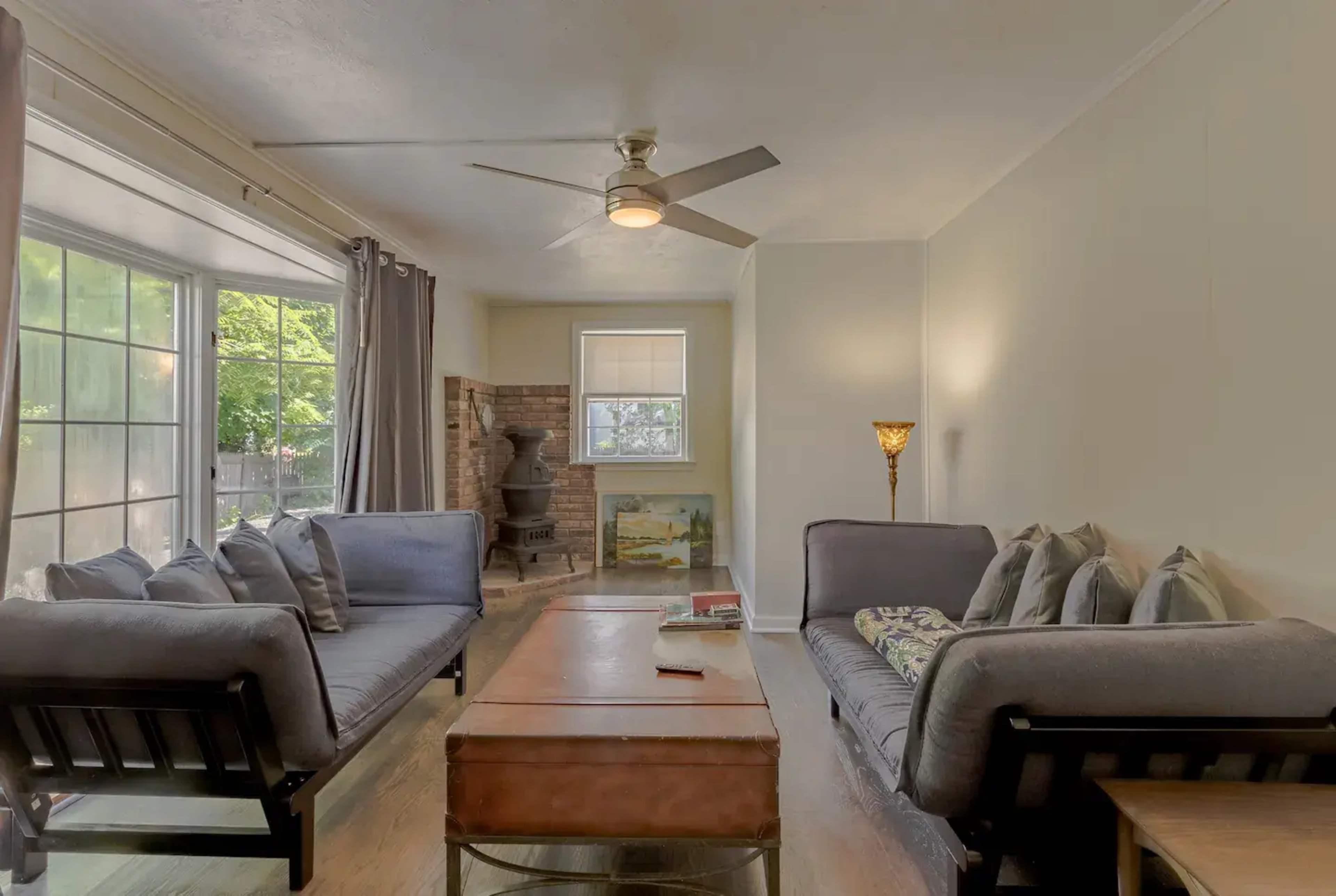 A living room with two gray sofas facing a wooden coffee table, a ceiling fan above, and a window letting in natural light.