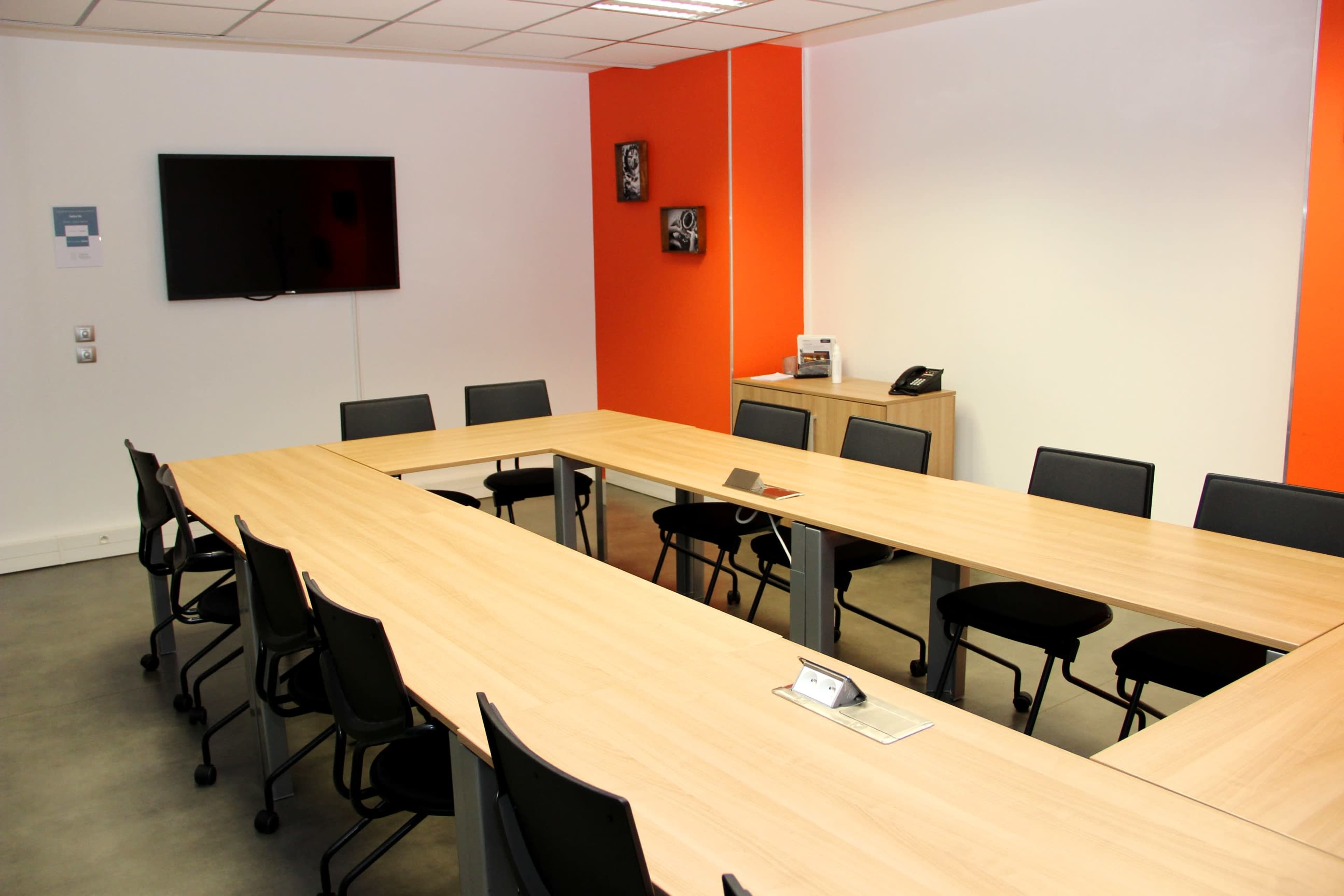 A rectangular conference room features a large wooden table surrounded by black chairs, equipped with a wall-mounted TV and a phone.