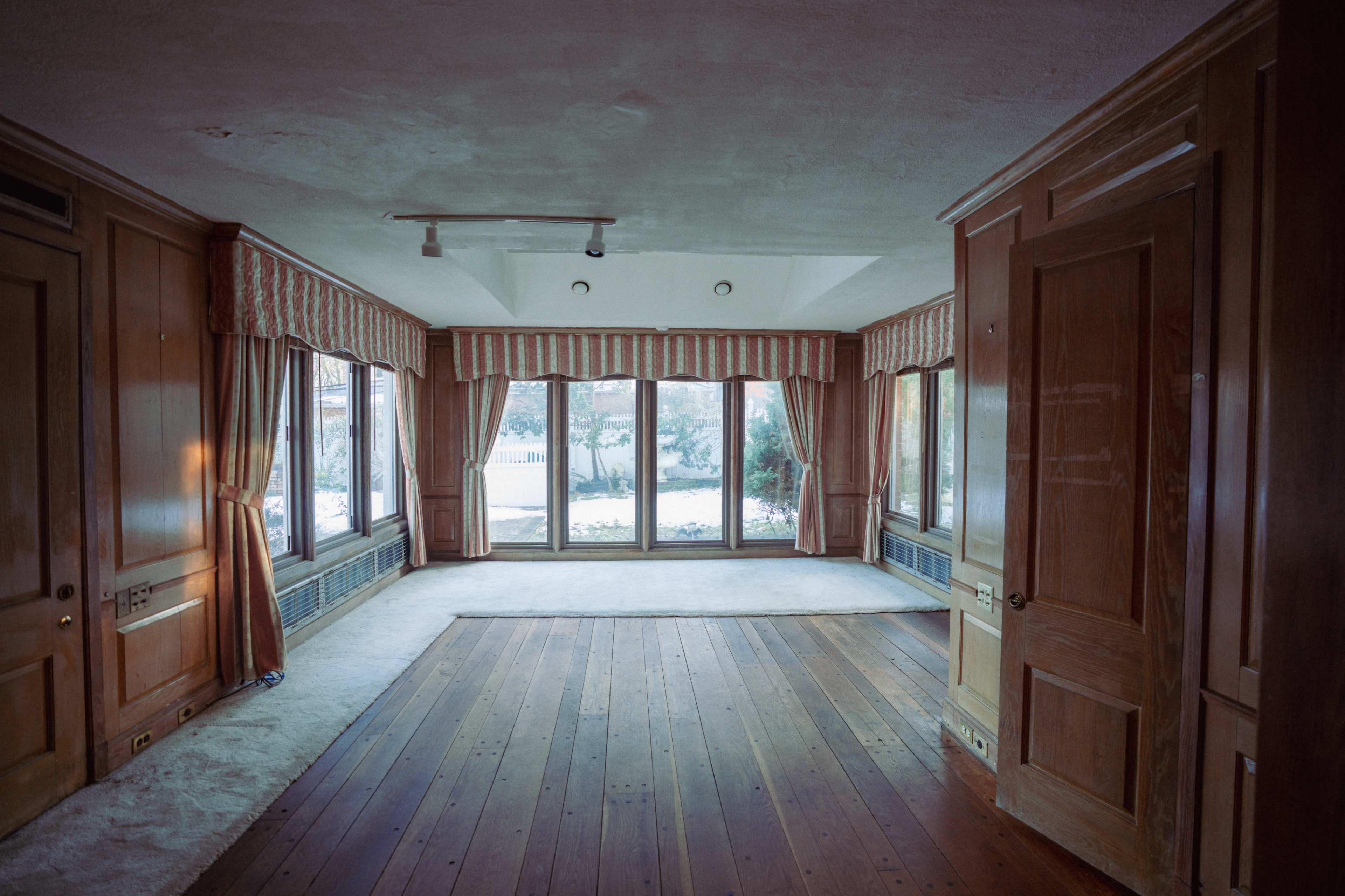 The image shows an empty room with wooden floors, large windows, and wooden paneling along the walls.