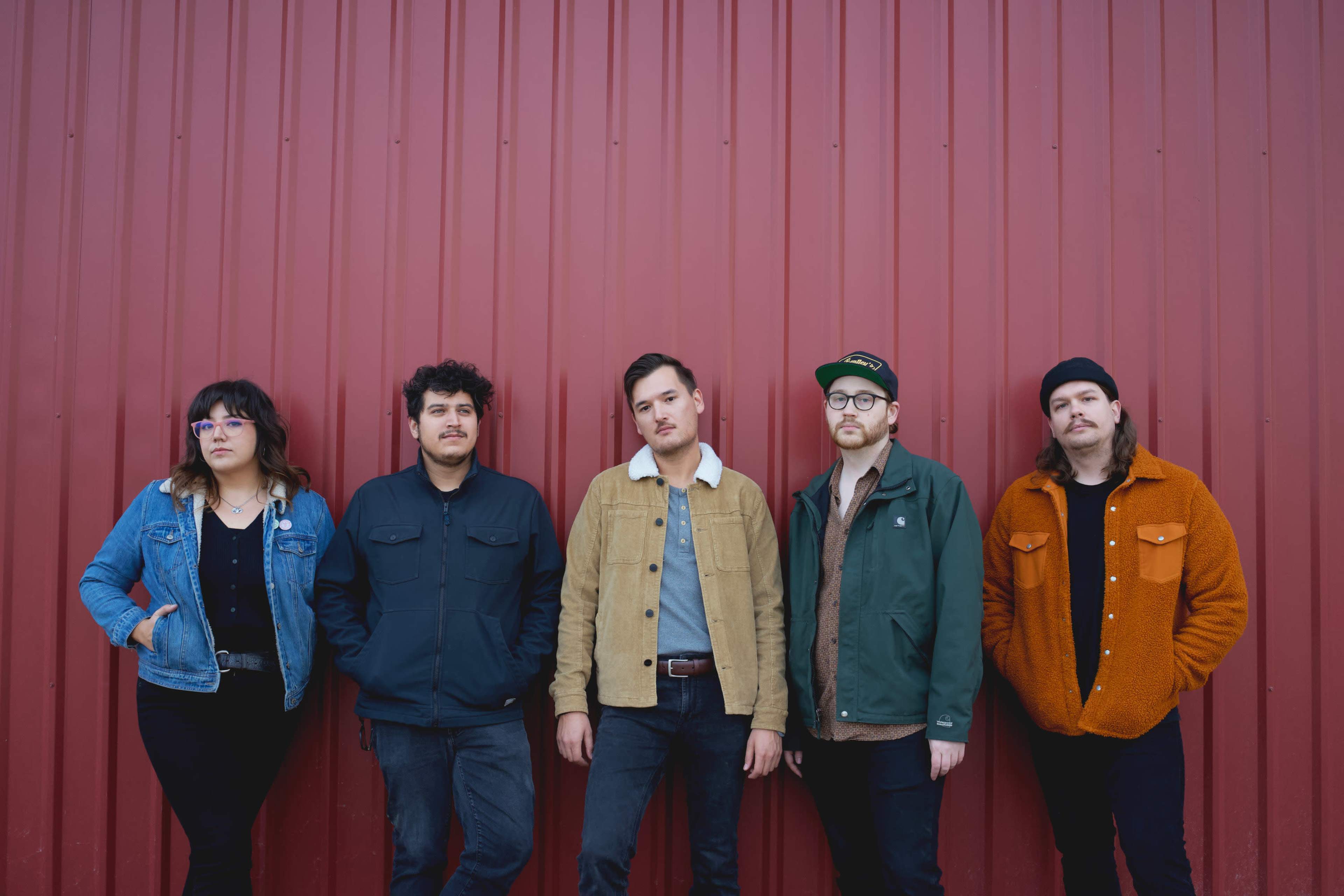 Five individuals stand in a line against a red corrugated metal wall, wearing various styles of jackets and shirts.