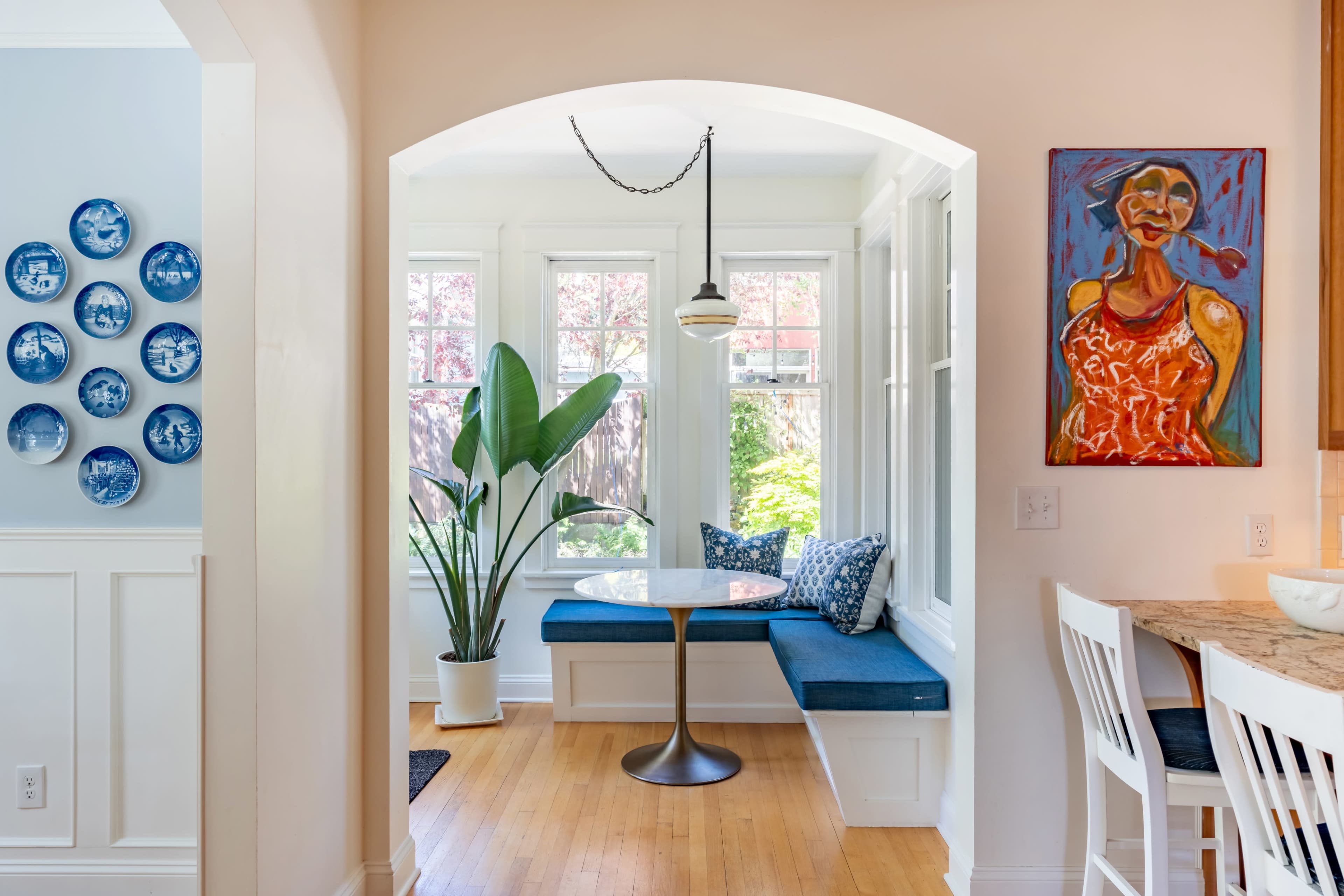 A bright dining area features a round table with a blue cushioned bench, framed by windows and decorated with a large painting and blue plates on the wall.