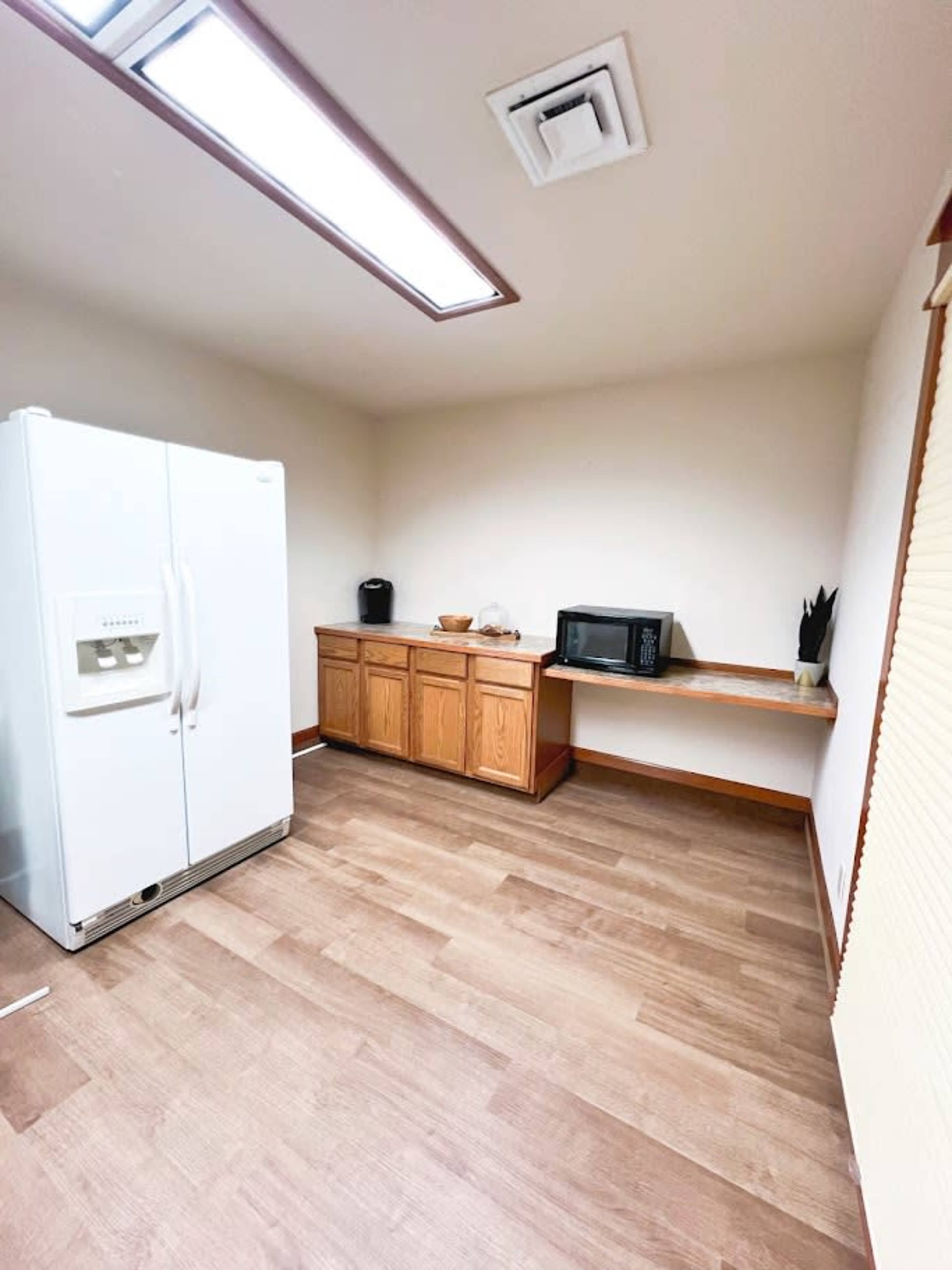The image shows a kitchen area with a white refrigerator, a microwave, and wooden cabinetry, featuring a wooden shelf and laminate flooring.