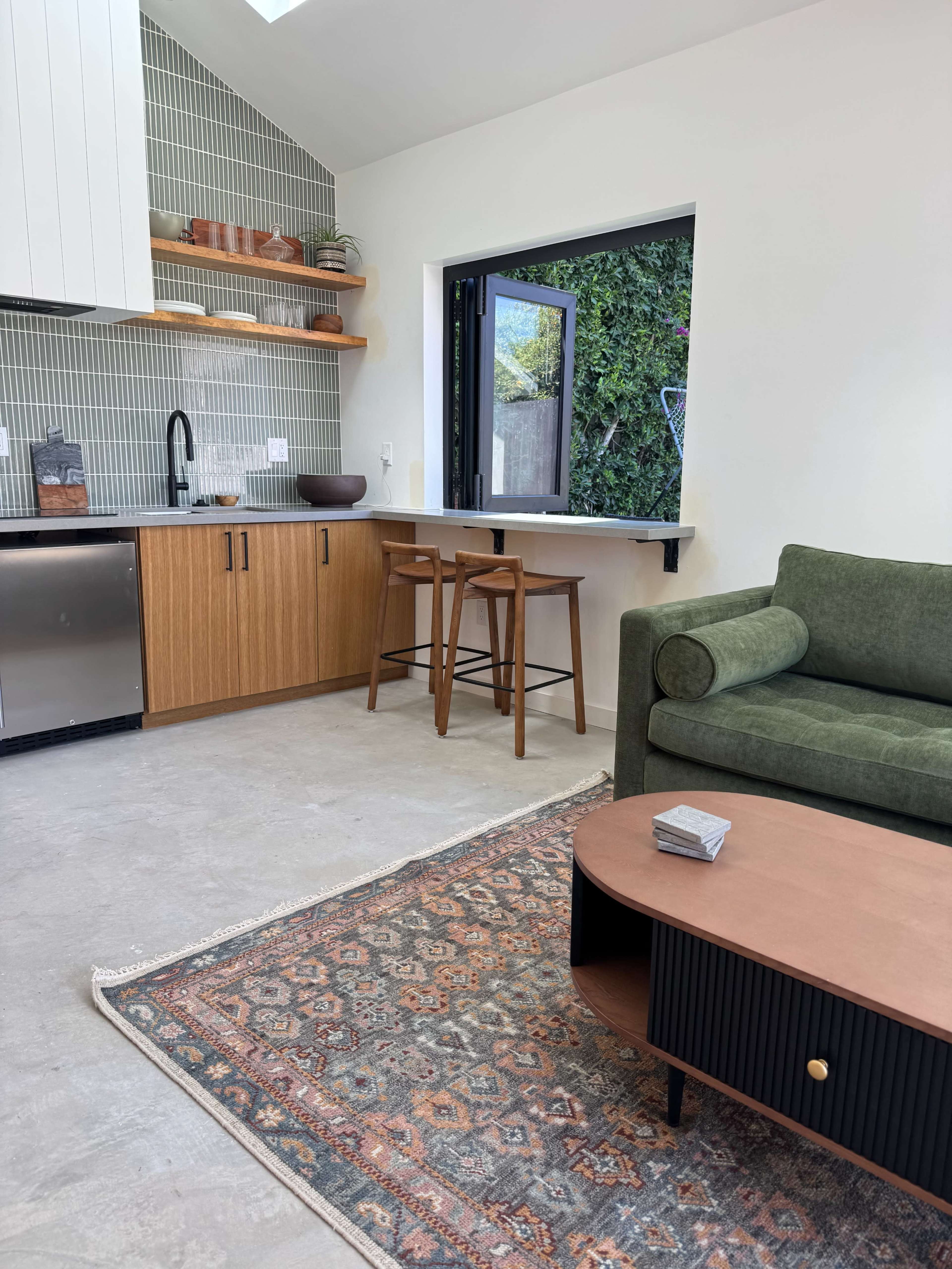 The image shows a modern kitchen and living area with a green couch, a coffee table, and a window overlooking a garden.