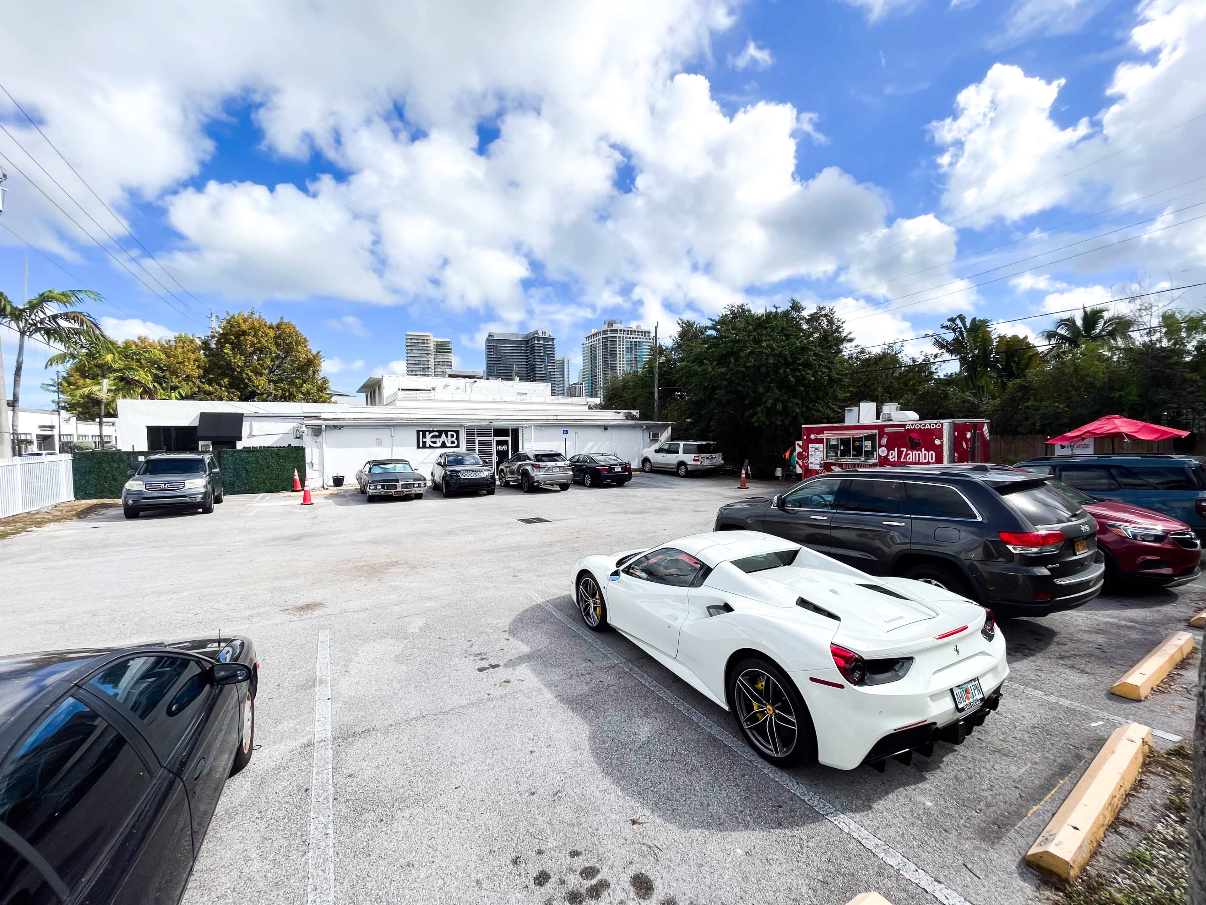 A white sports car is parked in a lot surrounded by several other vehicles, with a food truck and a city skyline visible in the background.