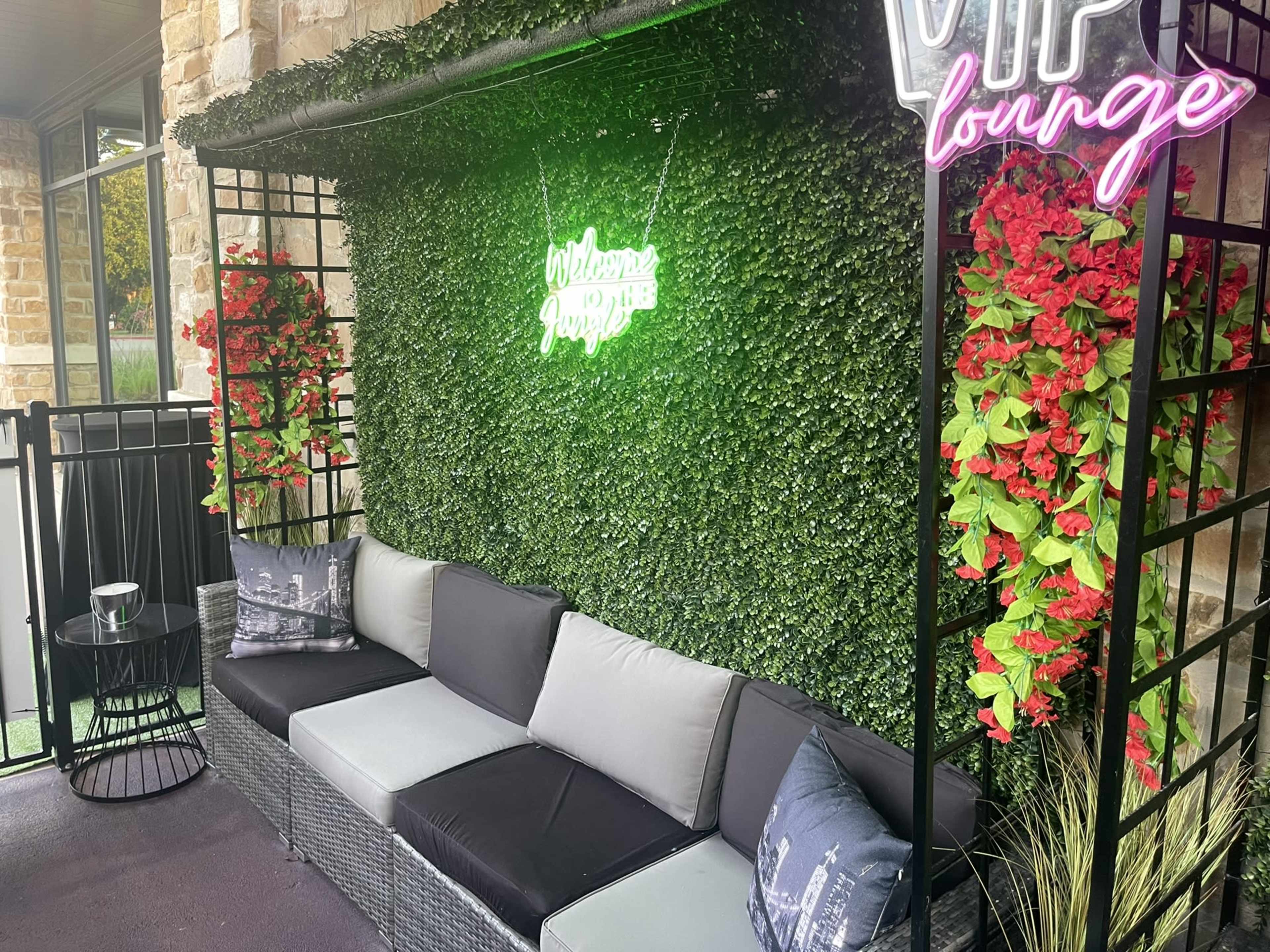 A VIP lounge area with a green wall of artificial foliage, vibrant red flowers, a modern couch, and a neon sign saying "Welcome to the VIP Lounge."