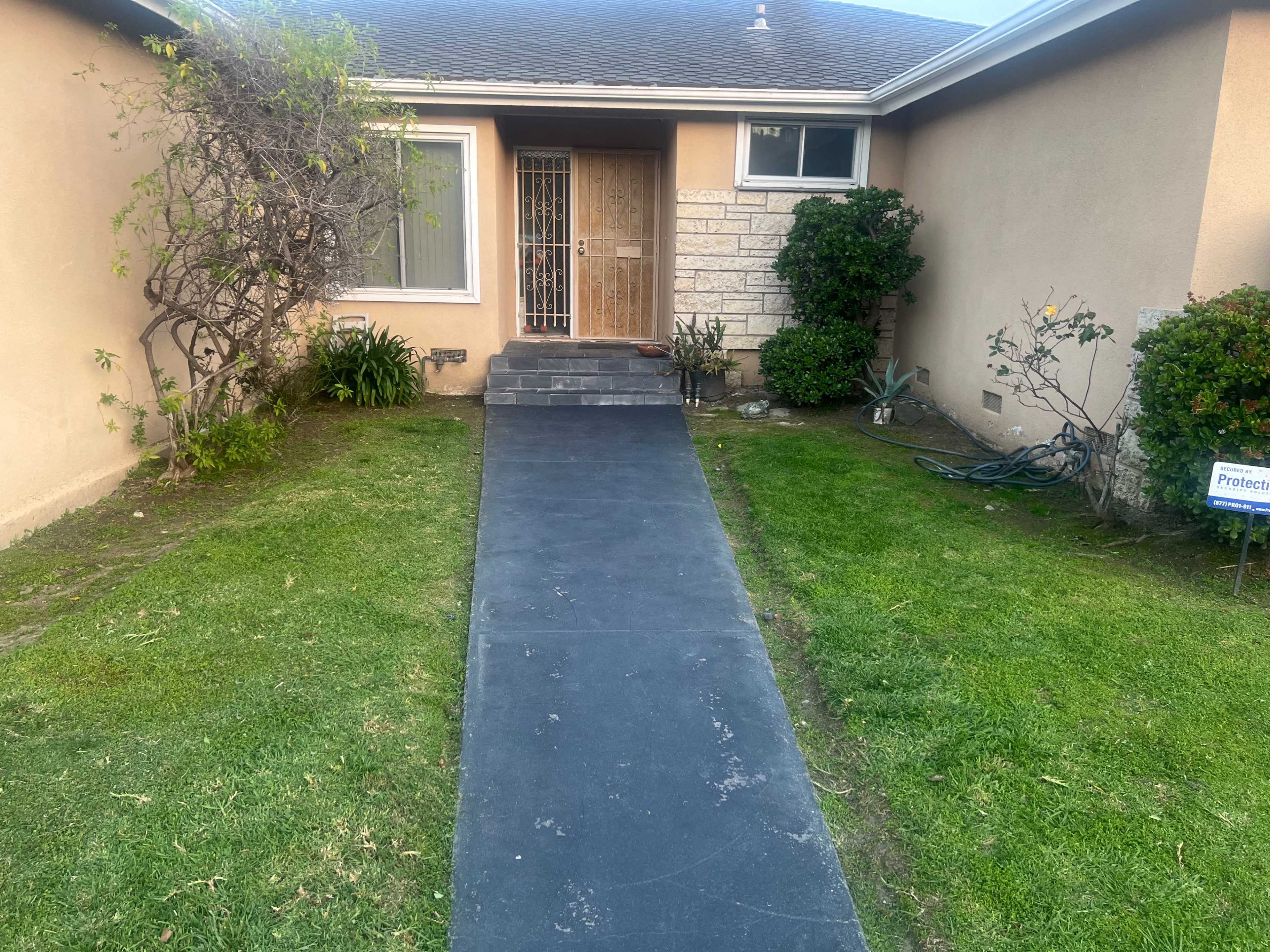 A pathway leading to a front door of a house, framed by grass and bushes on either side.