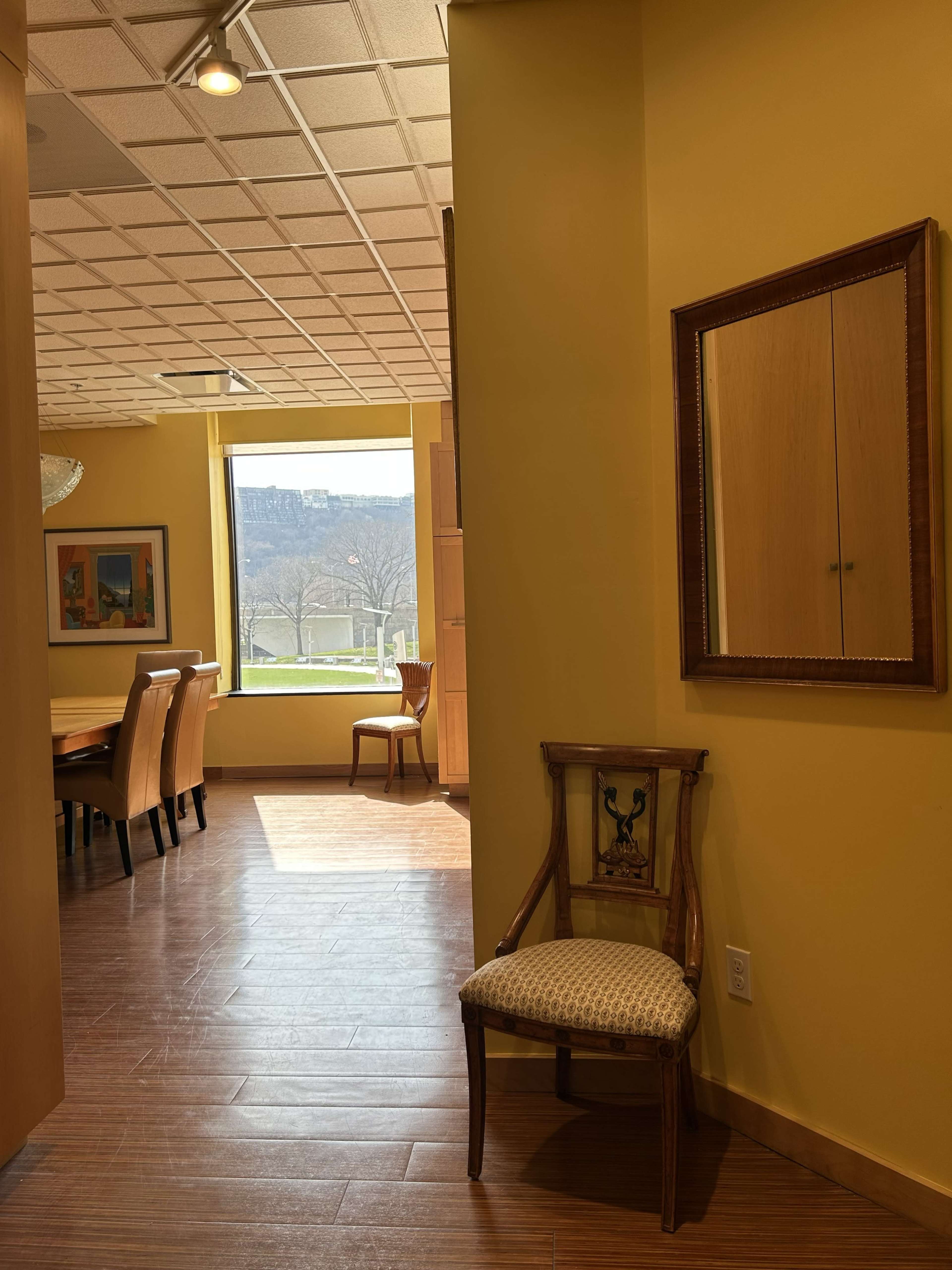 The image shows a brightly lit hallway leading to a dining area, with a chair and a large window showing a view of an outdoor landscape.