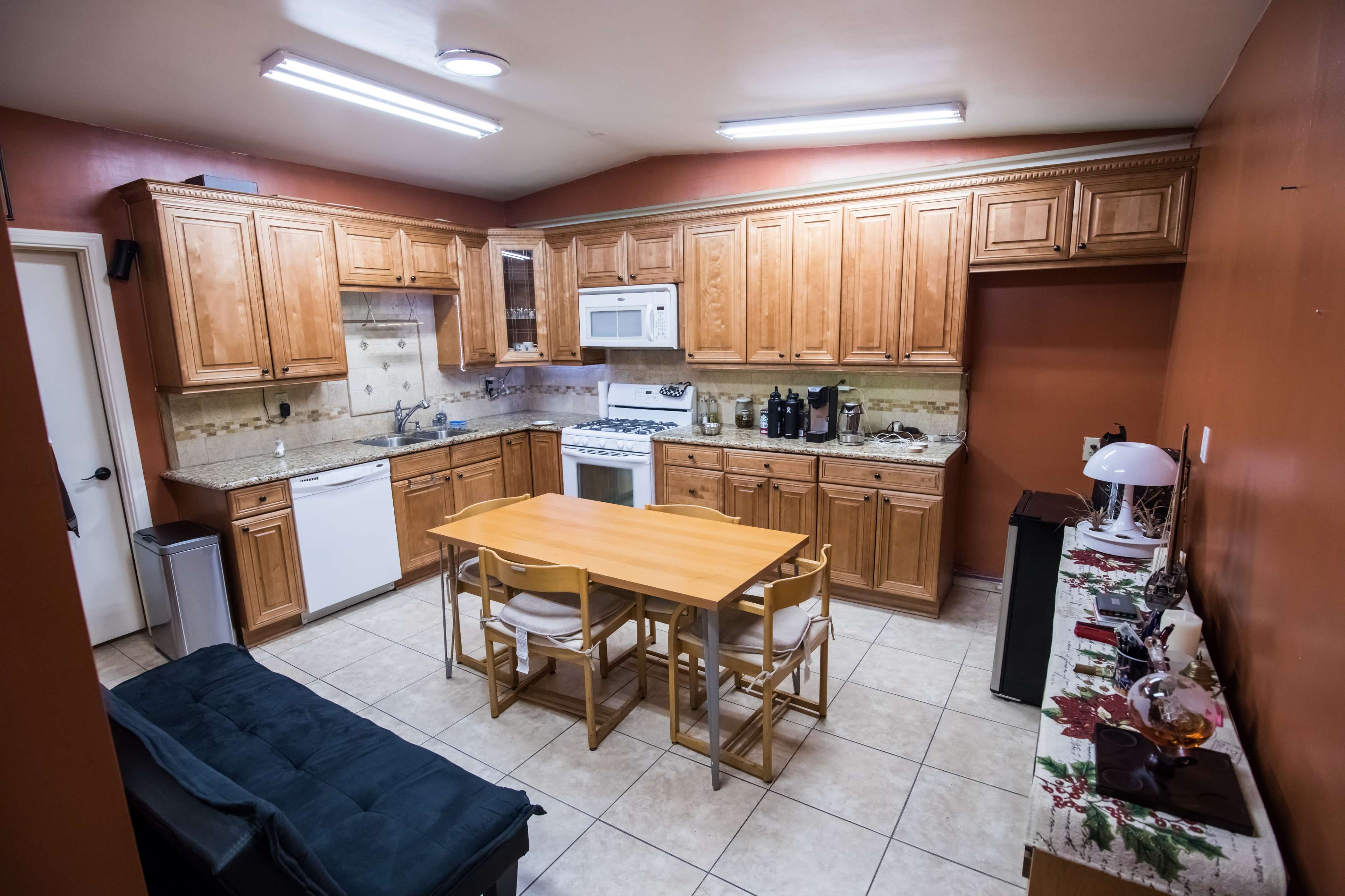 A kitchen with wooden cabinetry, a white fridge and microwave, a stove, a central dining table with chairs, and tiled flooring.