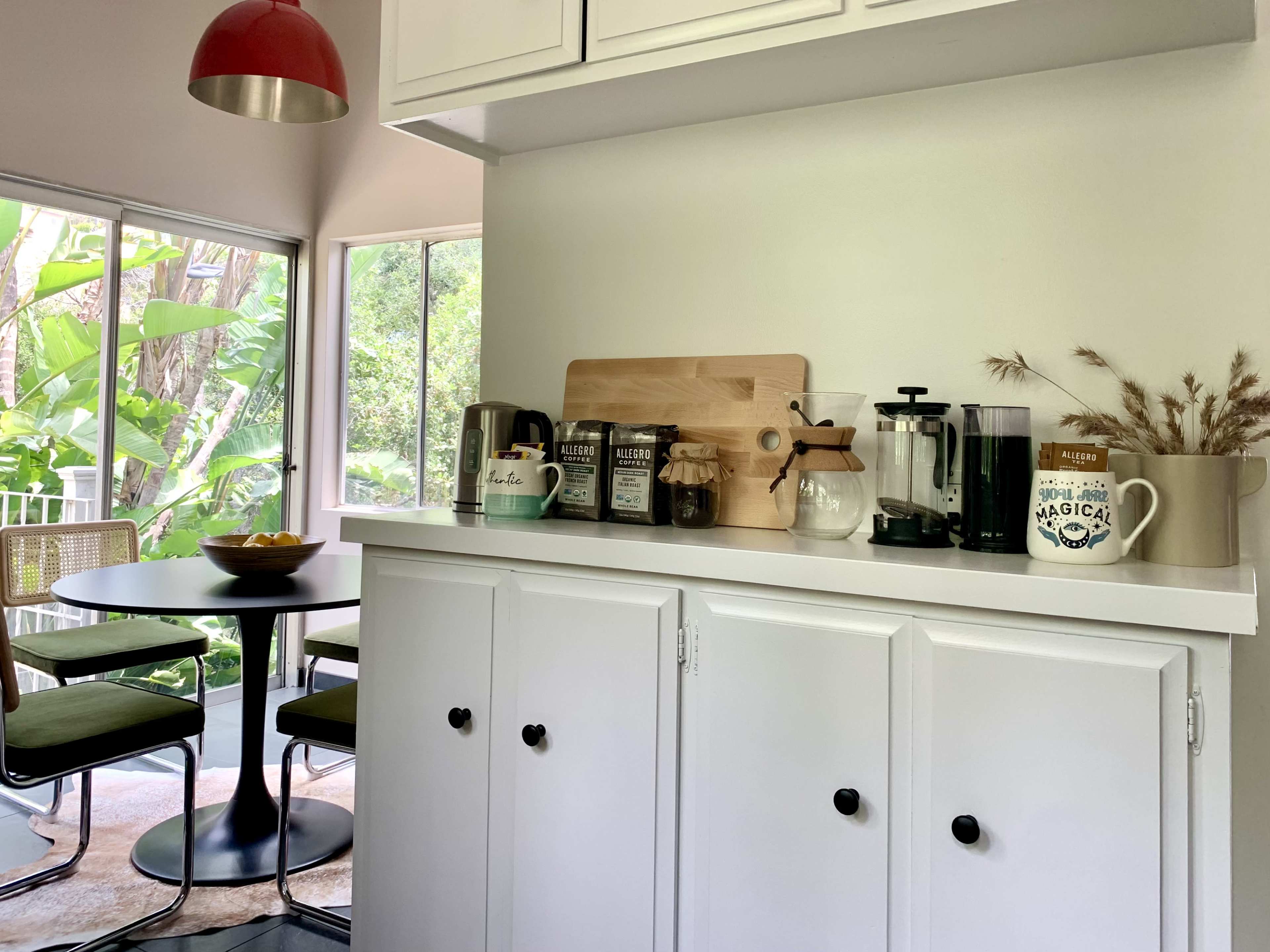 A bright kitchen features a small dining table, a coffee station with various appliances and utensils on a counter, and large windows overlooking greenery.