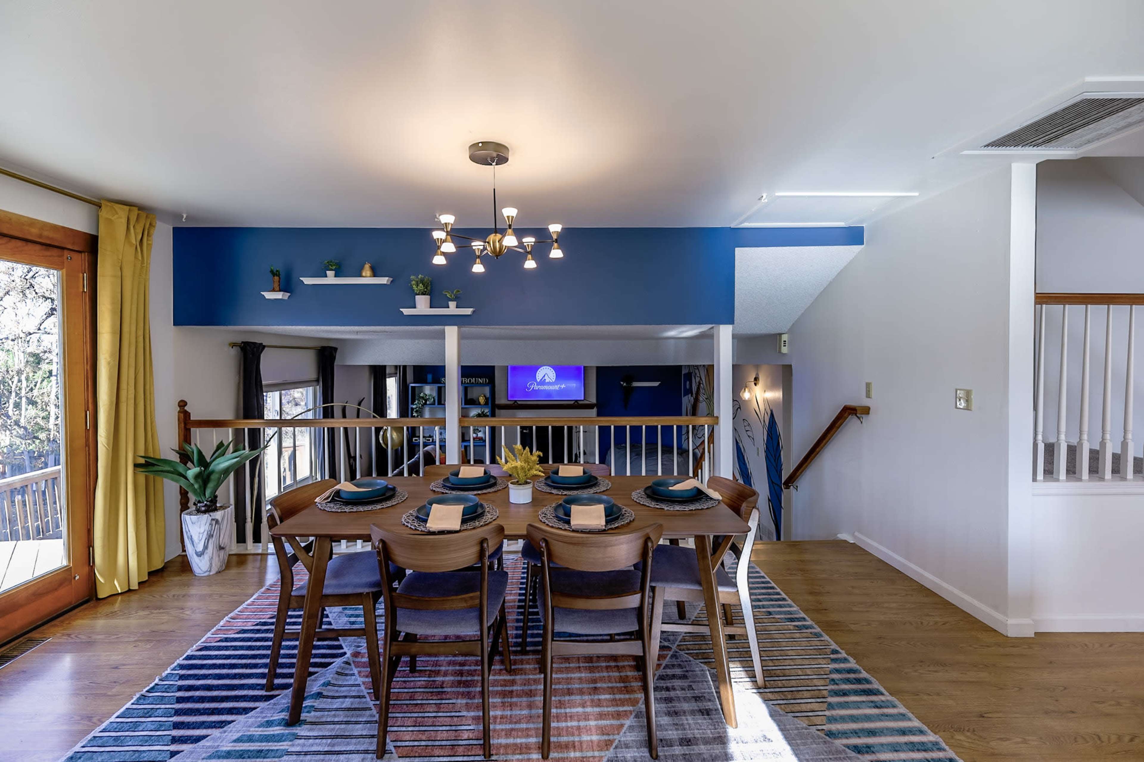 A dining area features a wooden table set for six, with a blue accent wall and a television visible in the background.