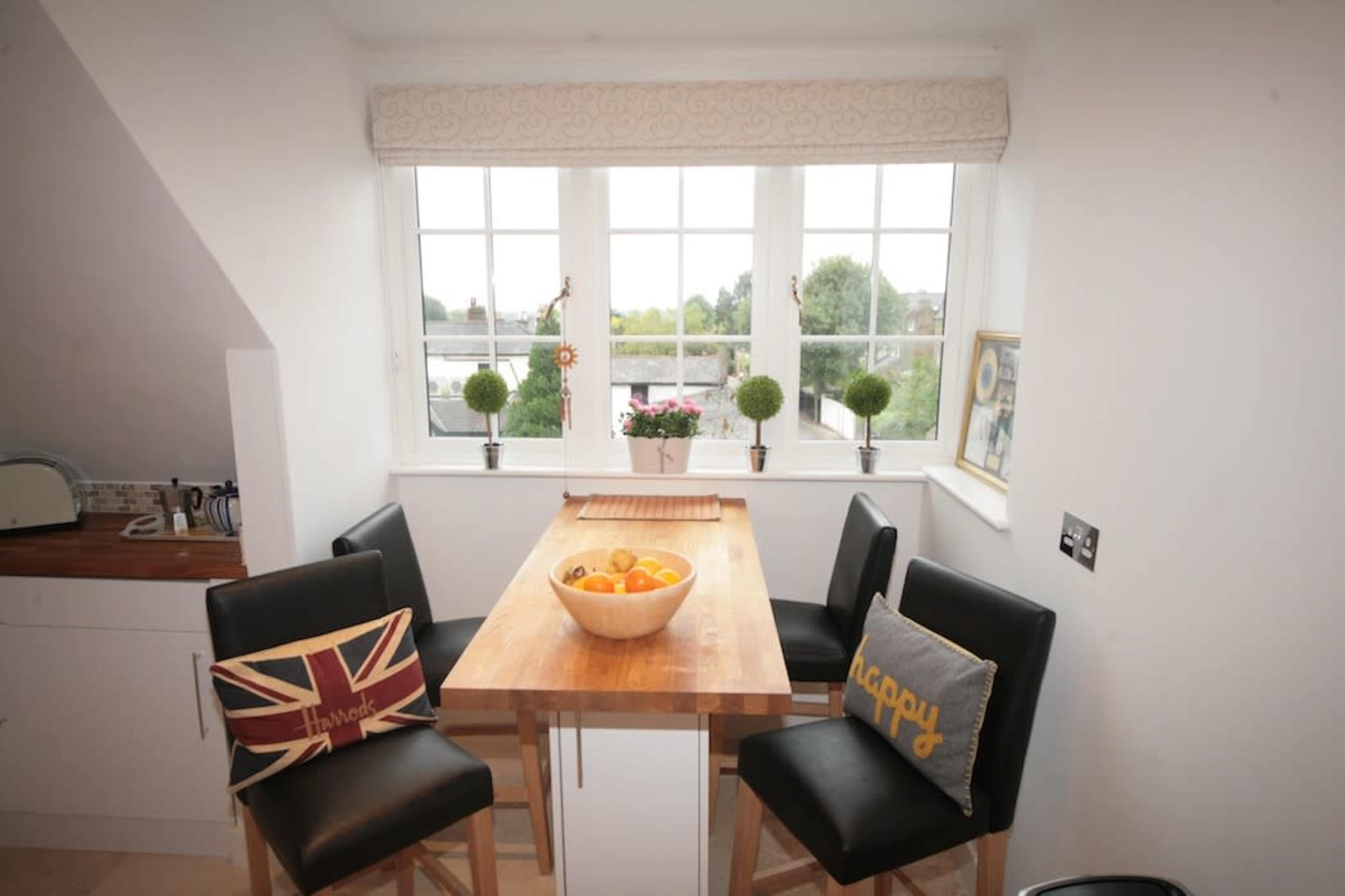 A small dining area features a wooden table with a bowl of fruit and four high-backed chairs near large window with potted plants.