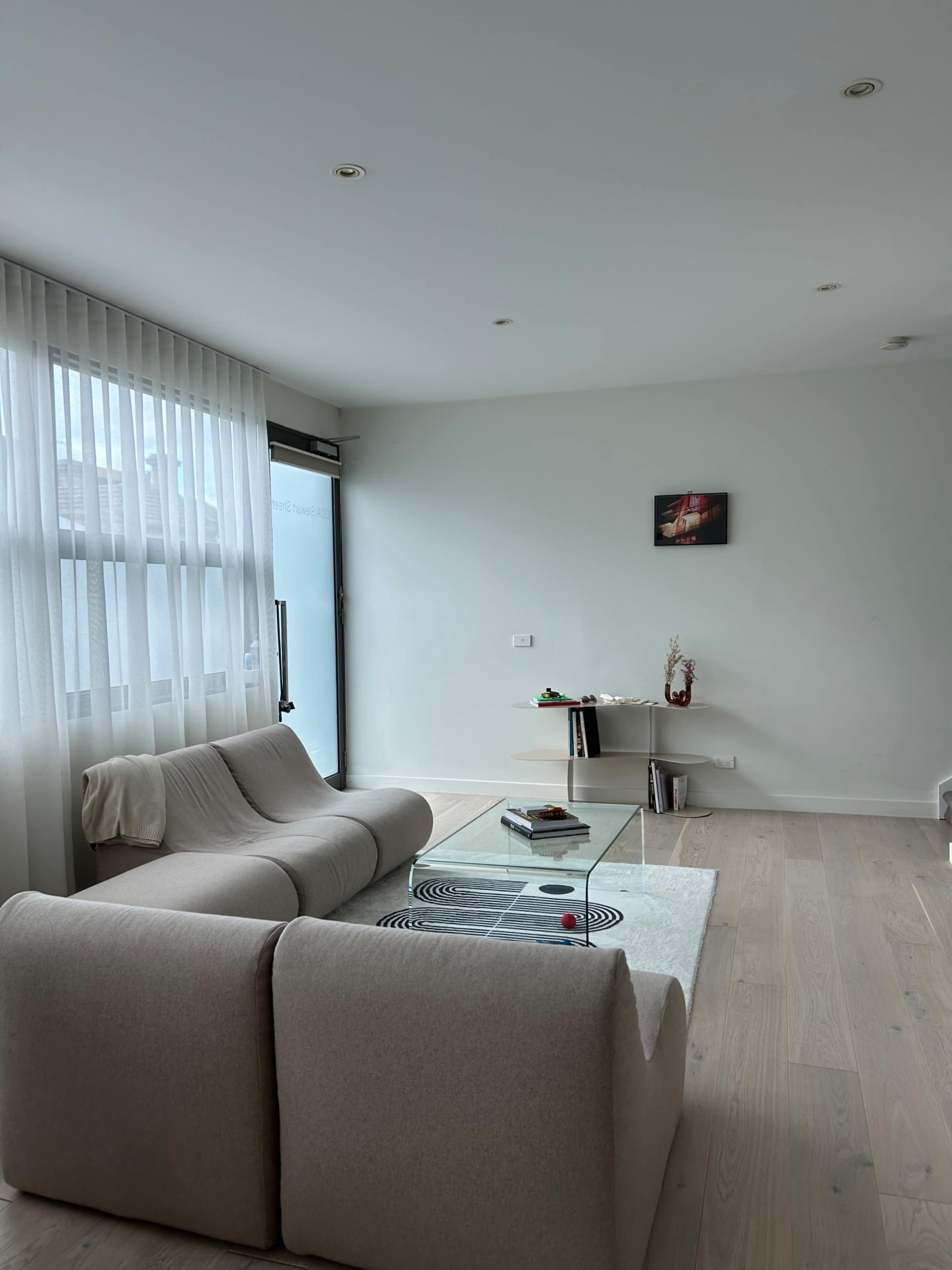 A modern living room with a light-colored sofa, a glass coffee table, and large windows draped with sheer curtains.