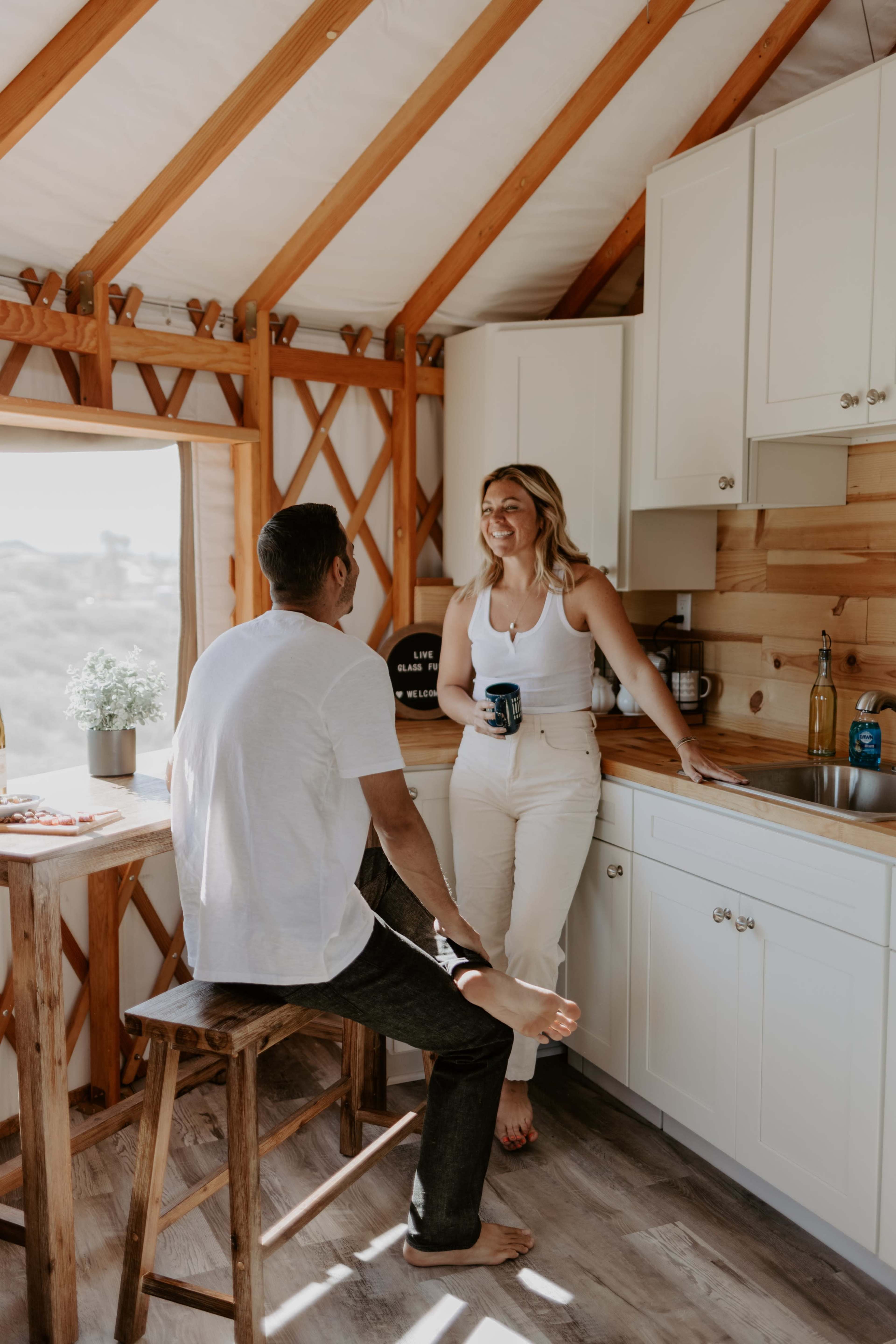 A man and a woman engage in conversation while standing in a rustic kitchen with wooden walls and white cabinetry.