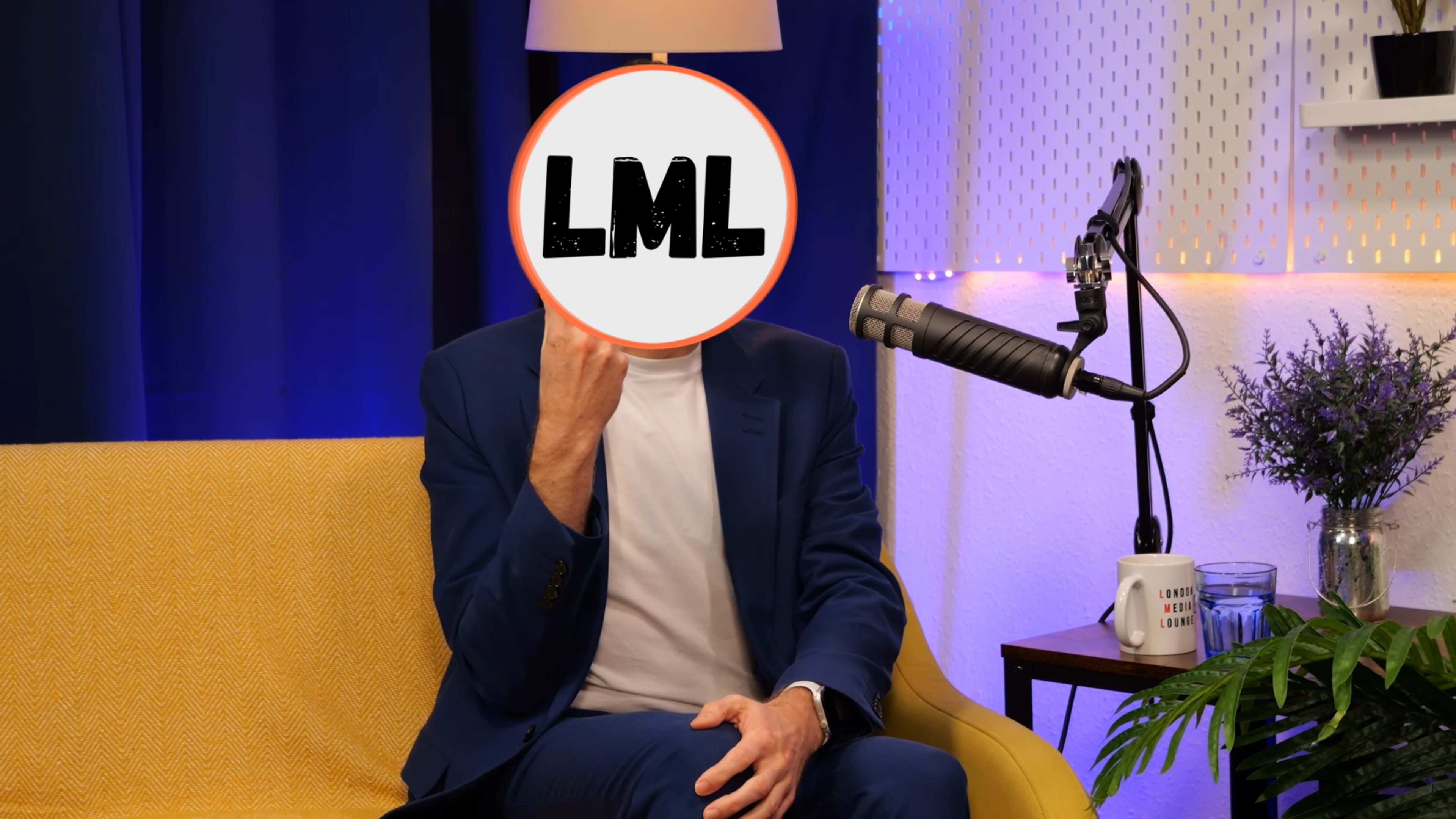 A person in a blue suit is sitting on a yellow couch in a studio, holding up a sign with the letters "LML" in a circle while a microphone and plant are visible nearby.