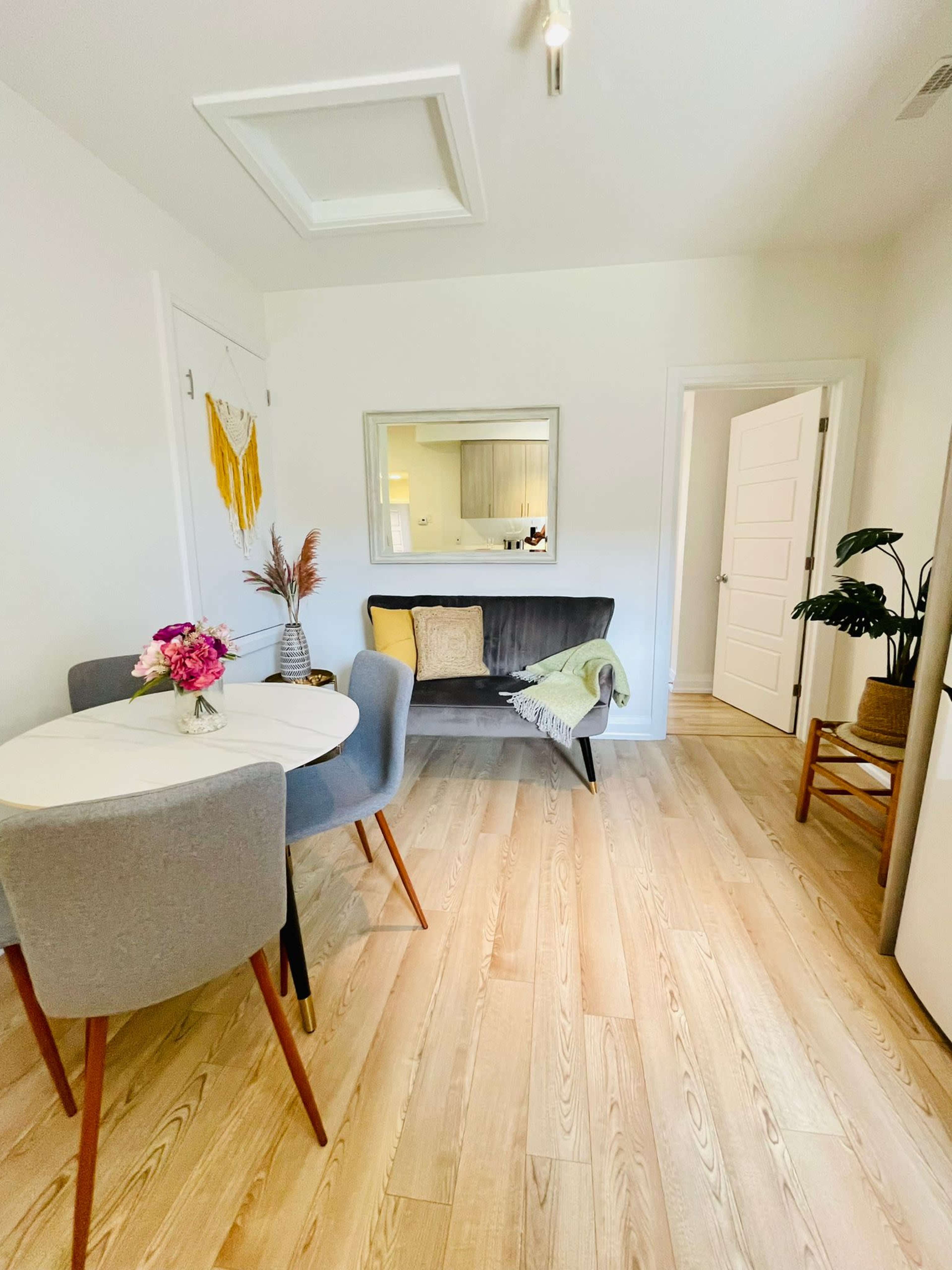 A small dining area with a round table and four chairs, a grey sofa against one wall, and light hardwood flooring.