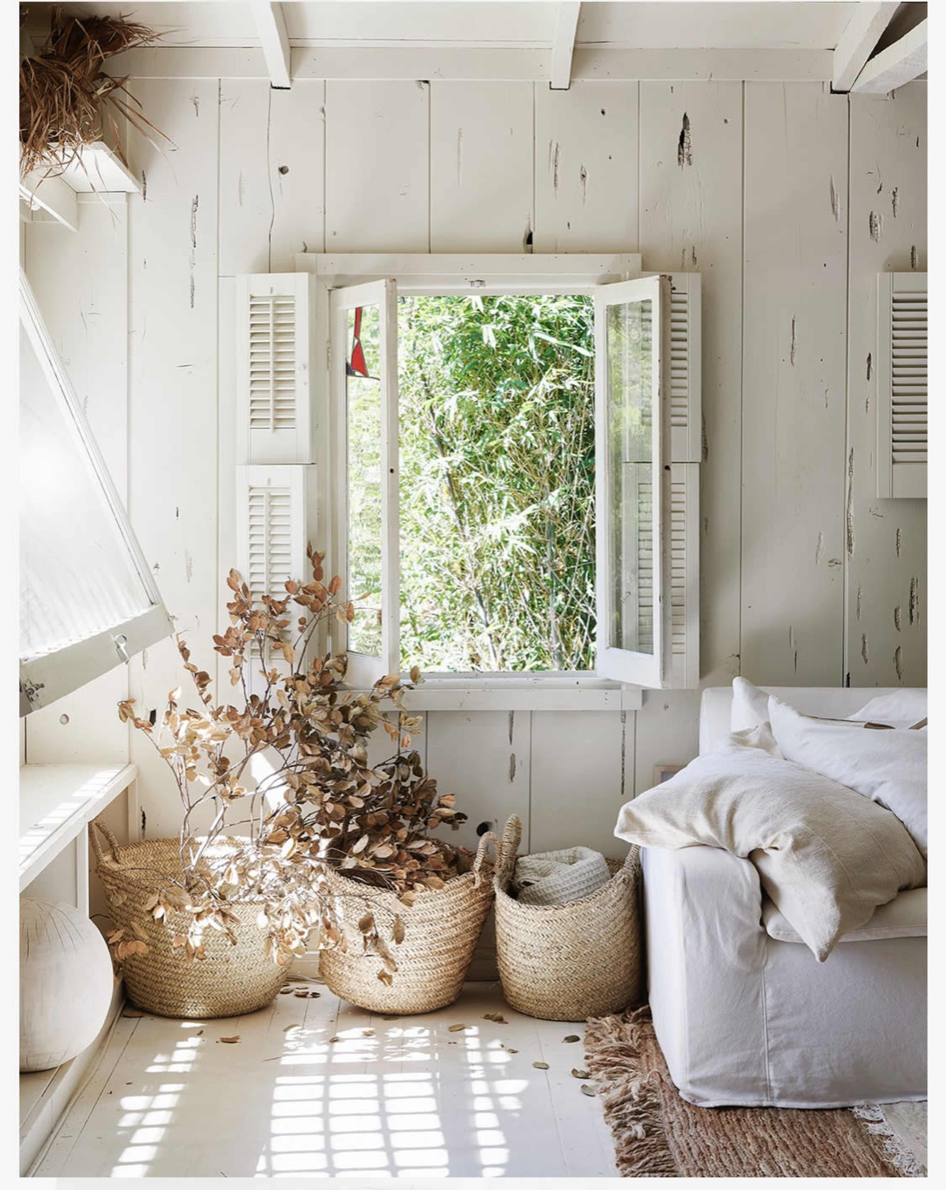 A sunlit room features open white shutters framing a view of greenery, with woven baskets and a white sofa adorned with pillows nearby.