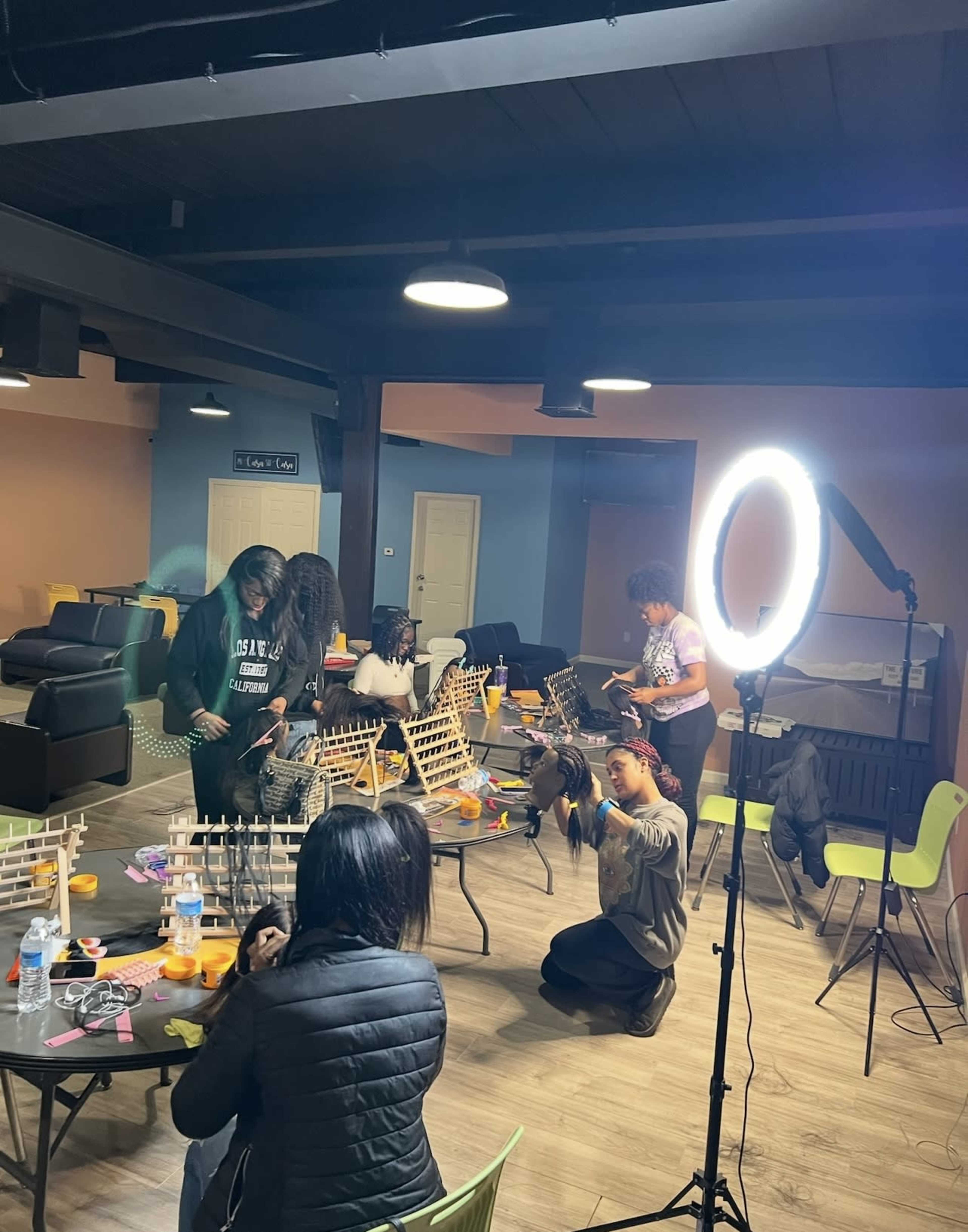 A group of individuals is engaged in a hair styling workshop in a brightly lit room equipped with seating and workspace.