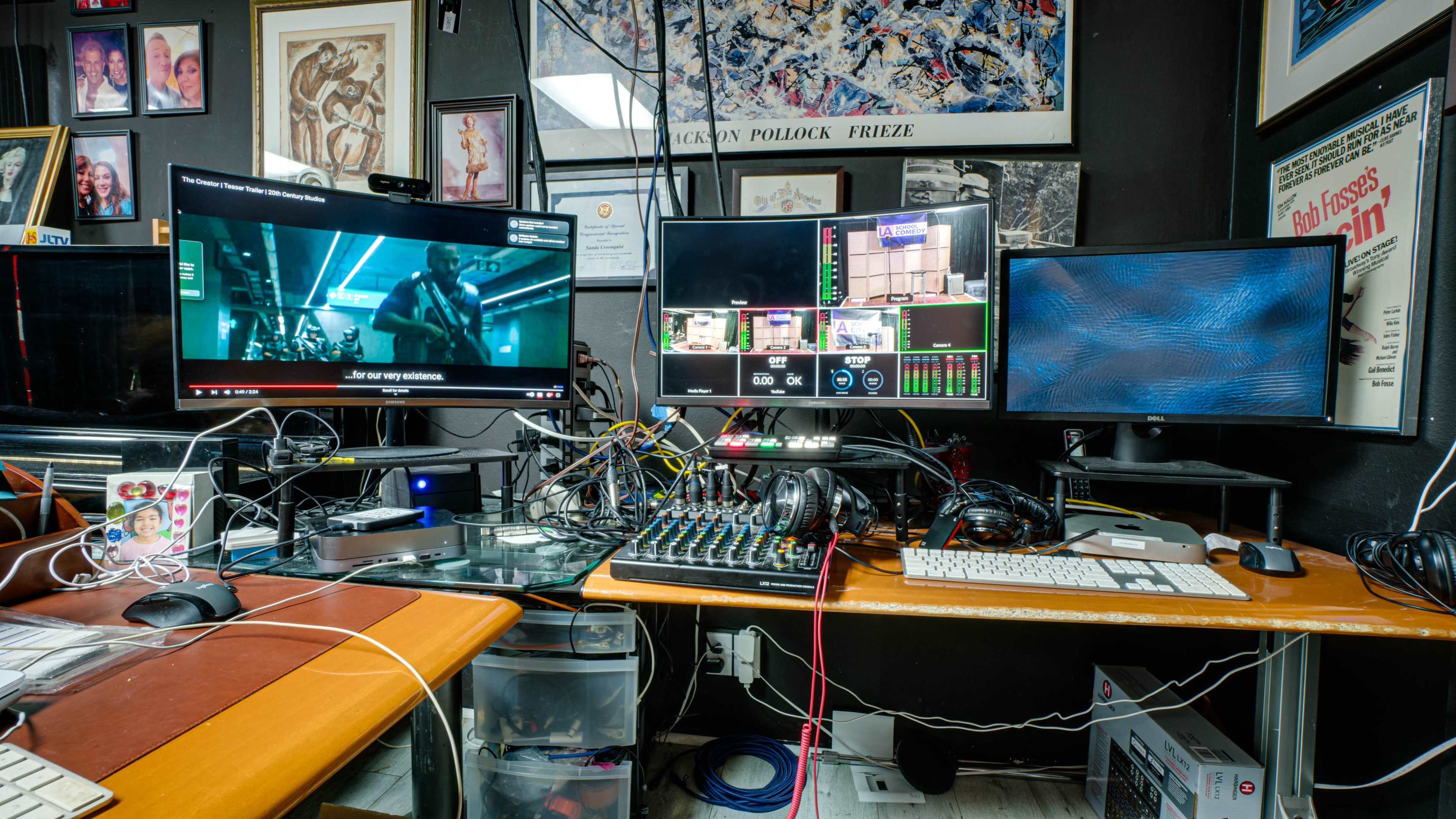 The image shows a cluttered workspace with multiple computer monitors displaying various video feeds, alongside a mixing console, keyboards, and tangled cables.