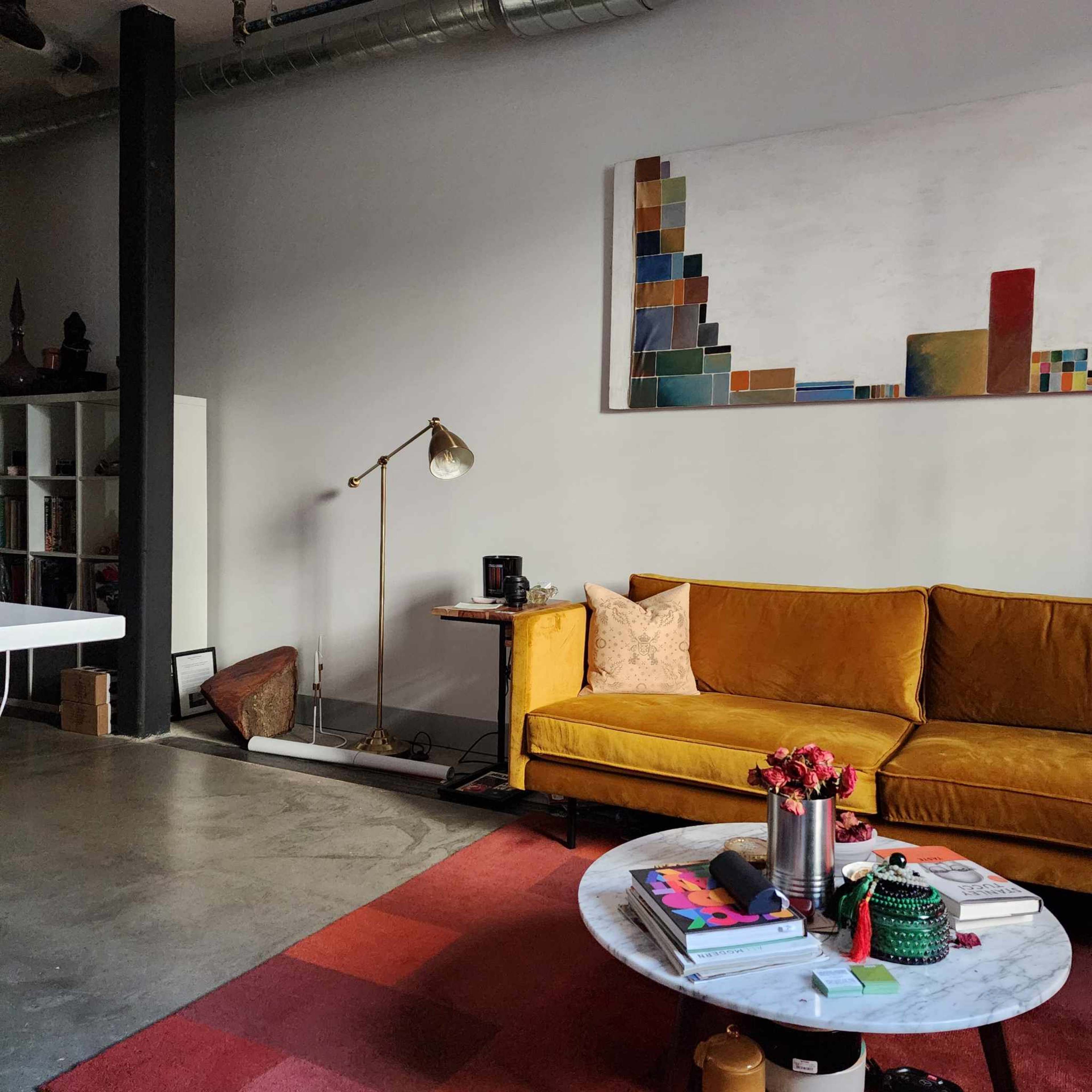 A modern living room features a yellow sofa, a marble coffee table with books, and wall art above a concrete floor.