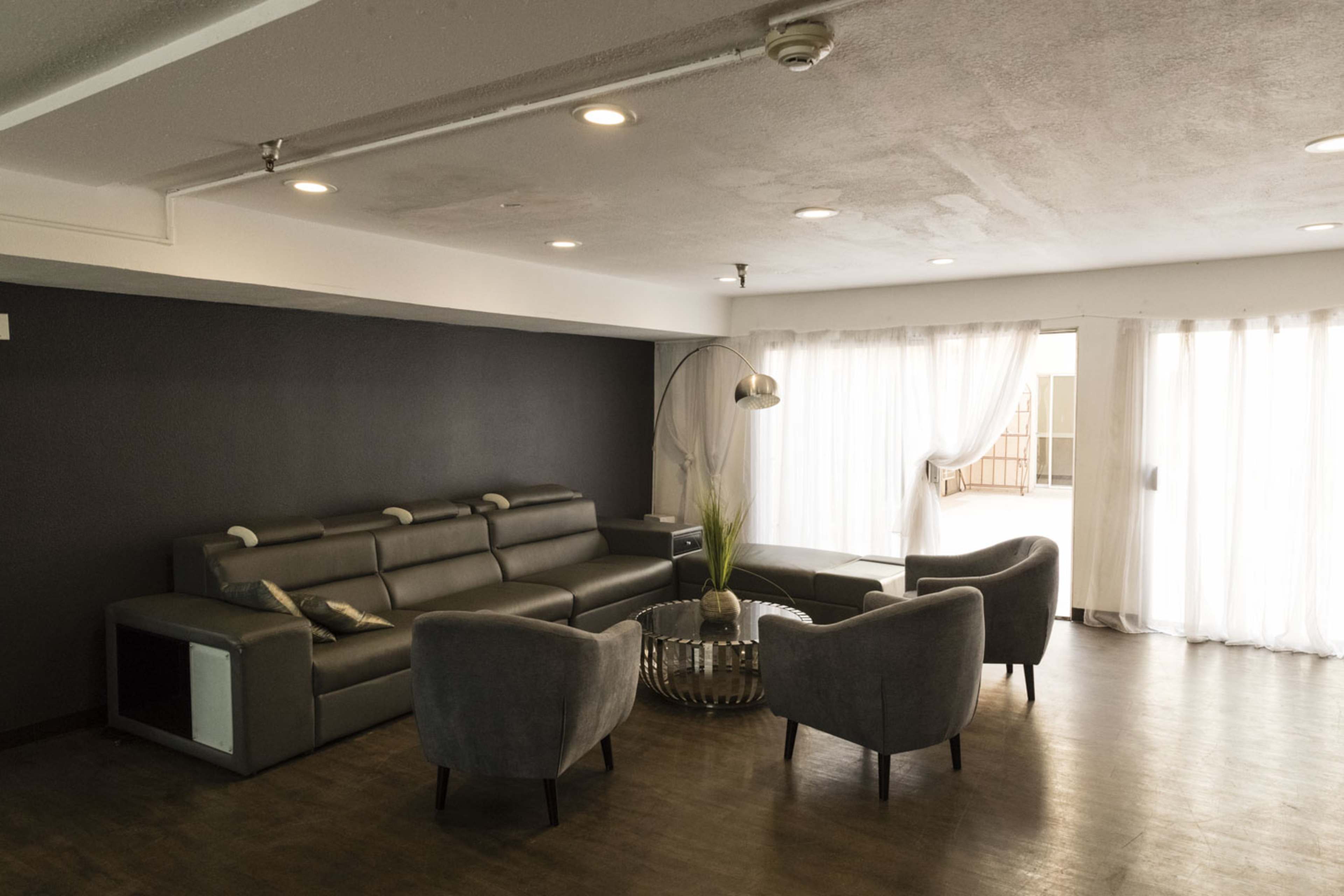 A modern living room features a gray leather sectional sofa, two upholstered chairs, a round coffee table, and large windows with sheer curtains.