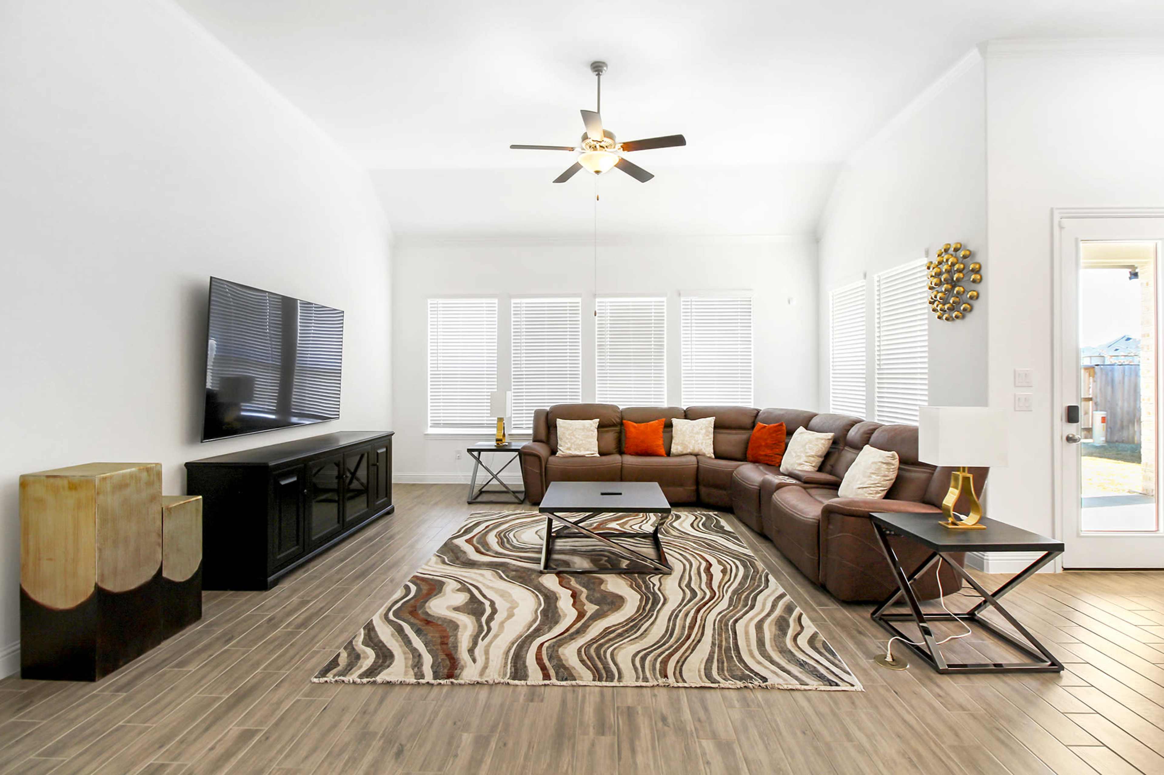 A modern living room featuring a brown sectional sofa, a coffee table, a large flat-screen TV on a console, and large windows allowing natural light.