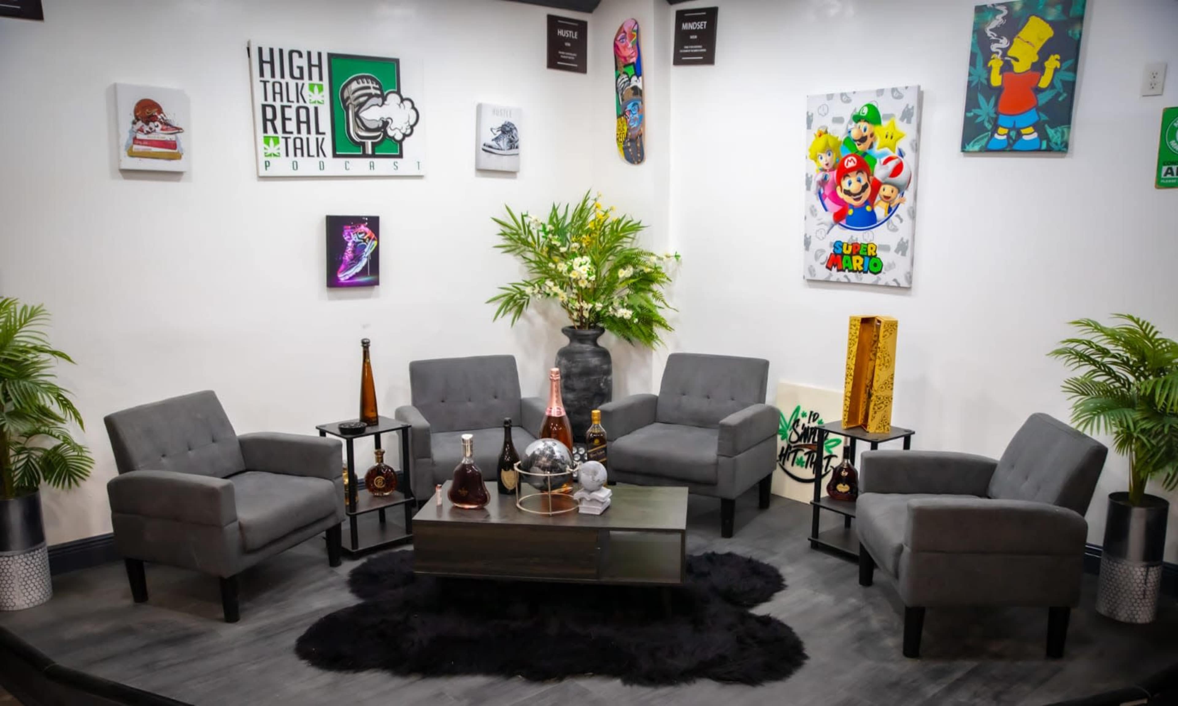 A cozy podcast recording studio with gray chairs arranged around a black coffee table, surrounded by vibrant artwork on the walls and green plants.