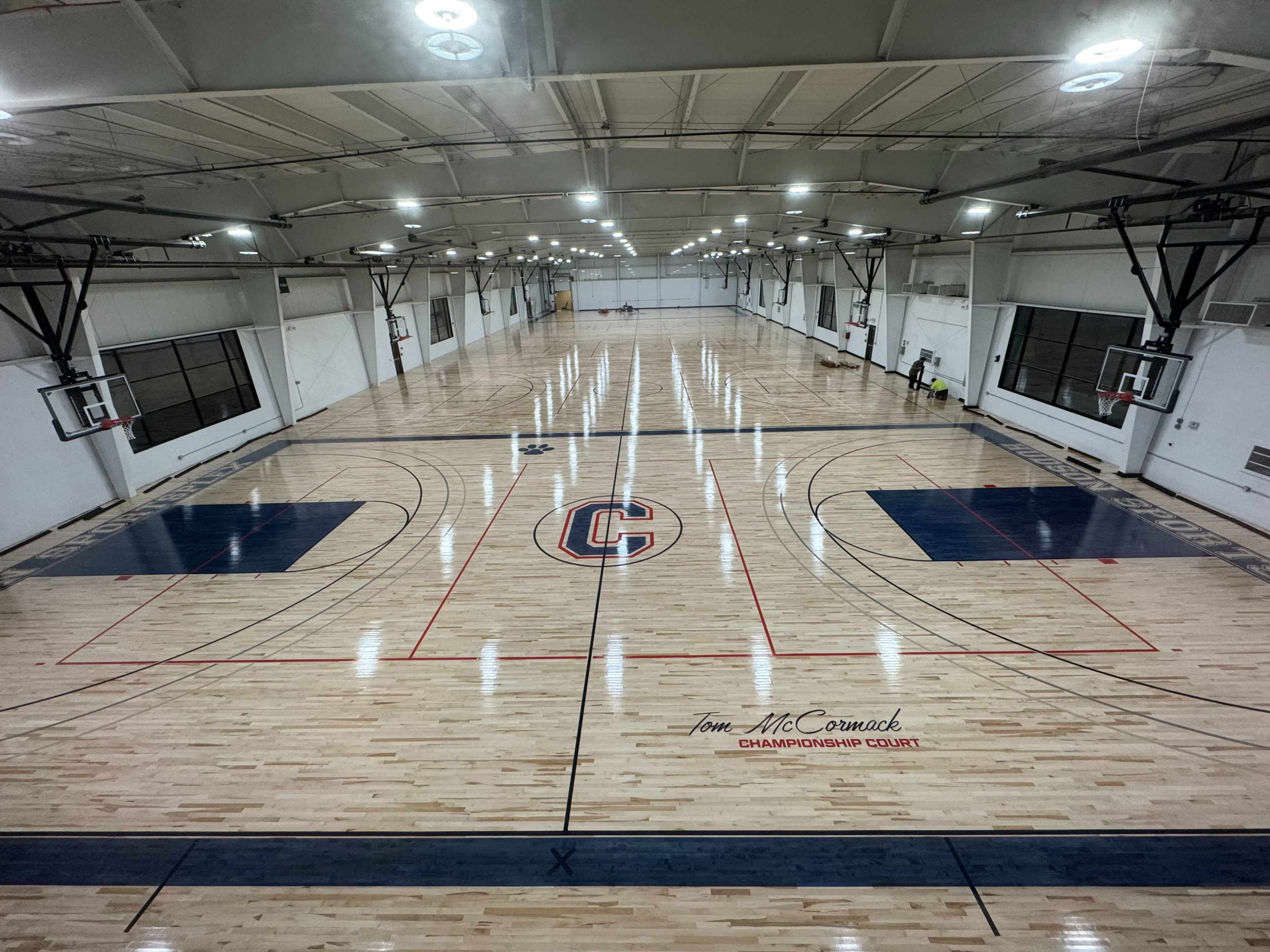 The image shows a spacious indoor basketball court with polished hardwood flooring, marked with red and blue lines, and multiple basketball hoops along the sides.