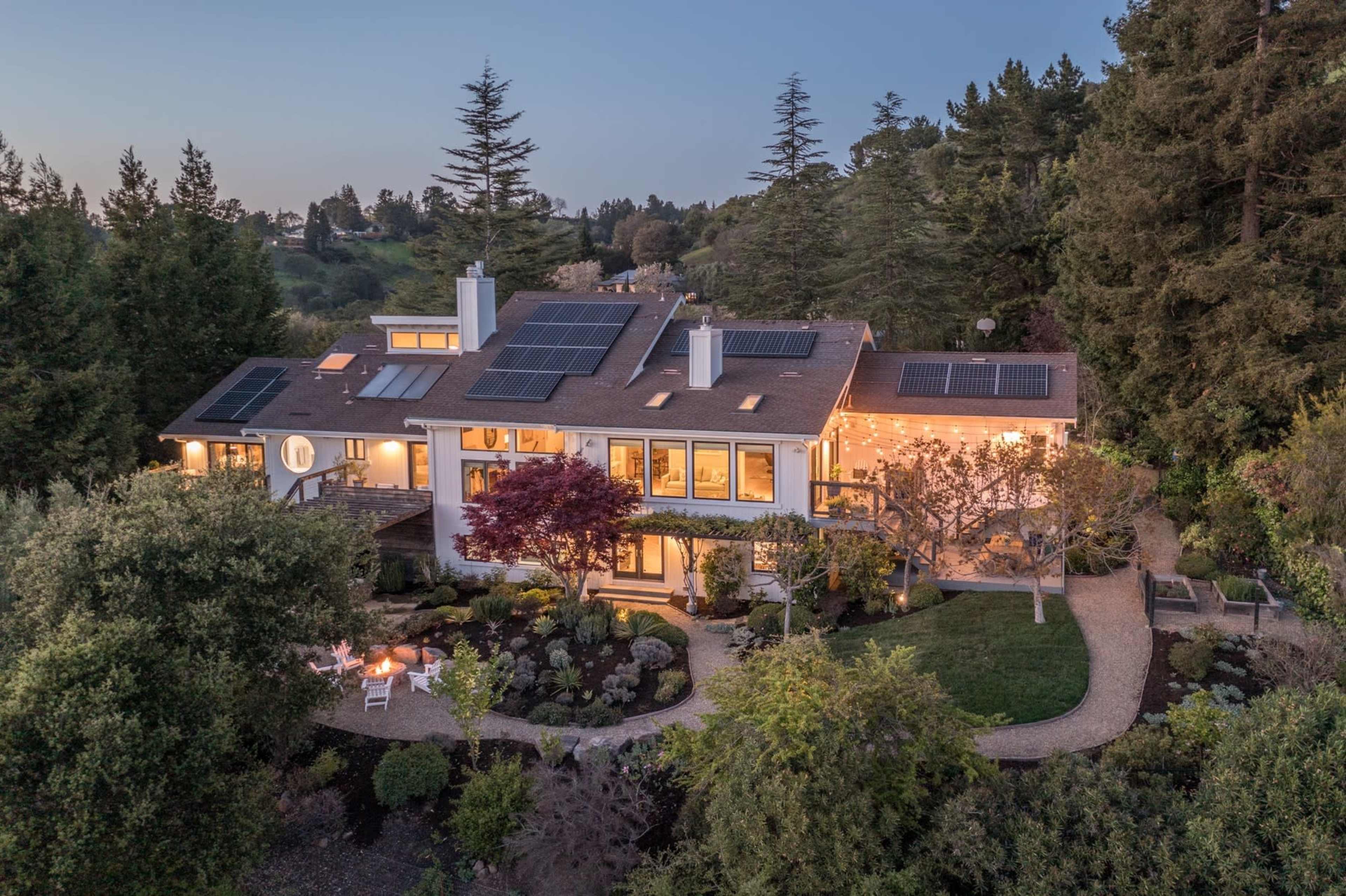 A modern house with solar panels on the roof, surrounded by landscaped gardens and trees at dusk.