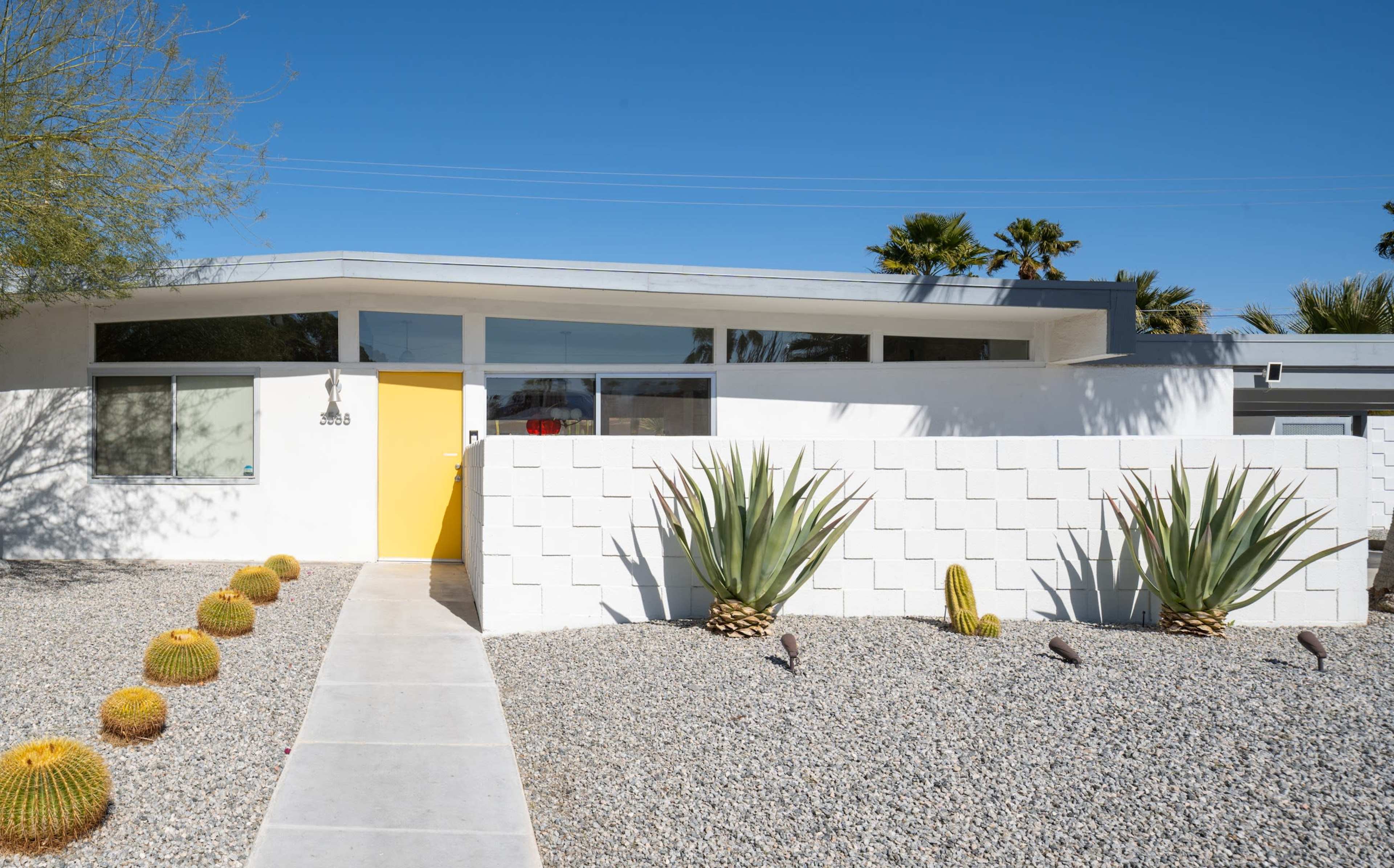 The image shows a modern white house with a bright yellow front door, surrounded by gravel landscaping and desert plants.