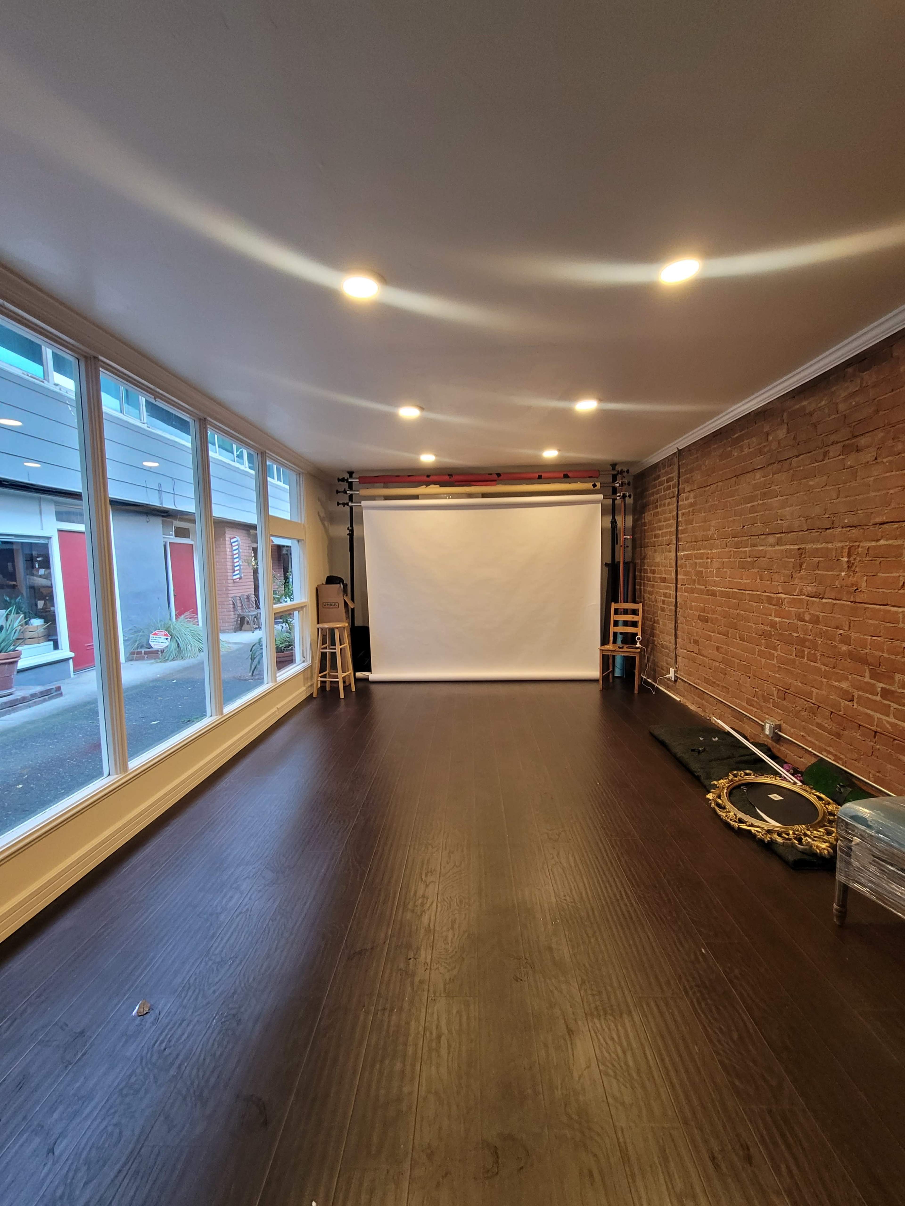The image shows an empty room with wooden floors, large windows, and a blank projection screen at the front.