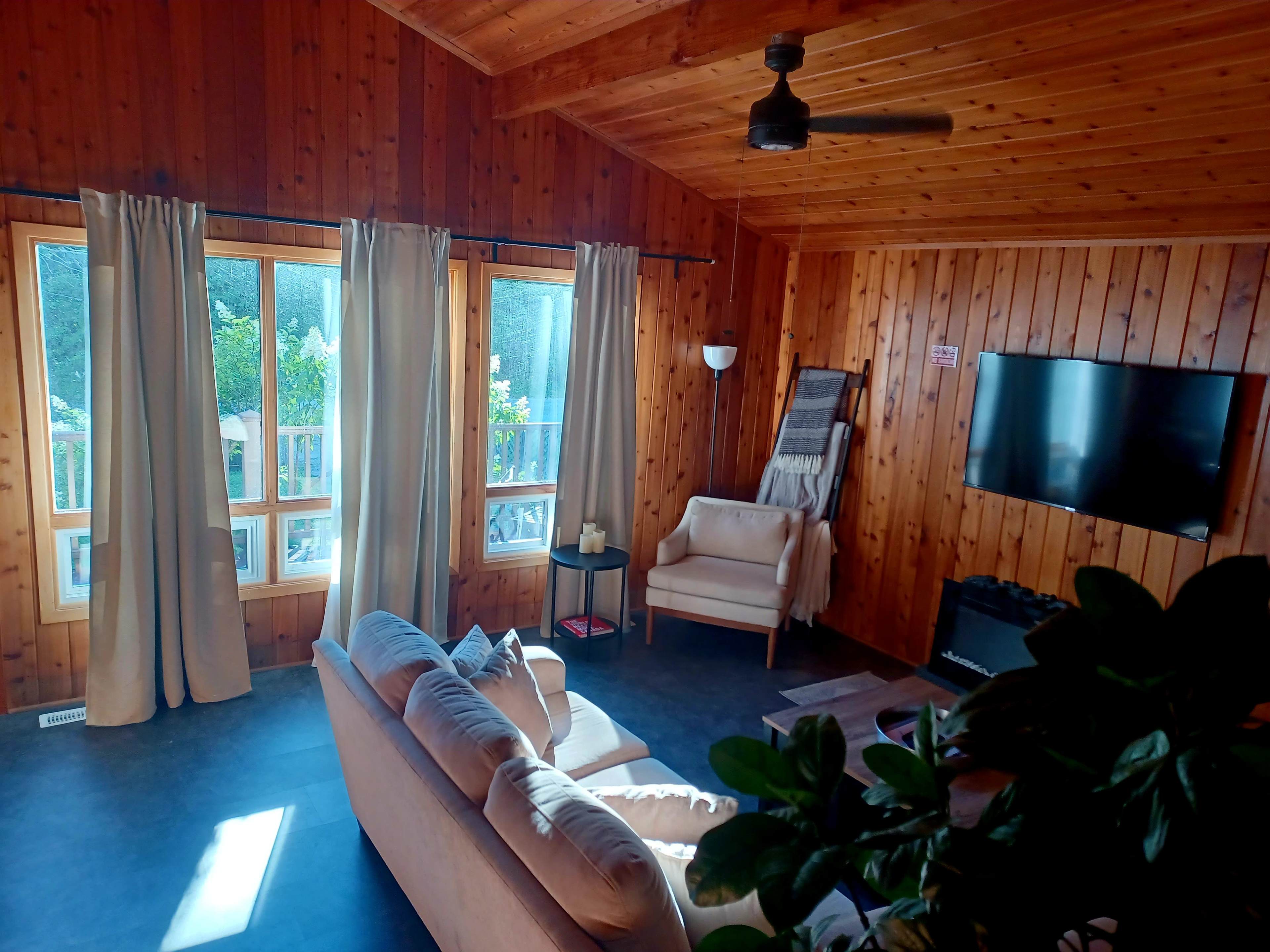 A cozy room with wooden walls, large windows, a light-colored sofa, and a television mounted on the wall.