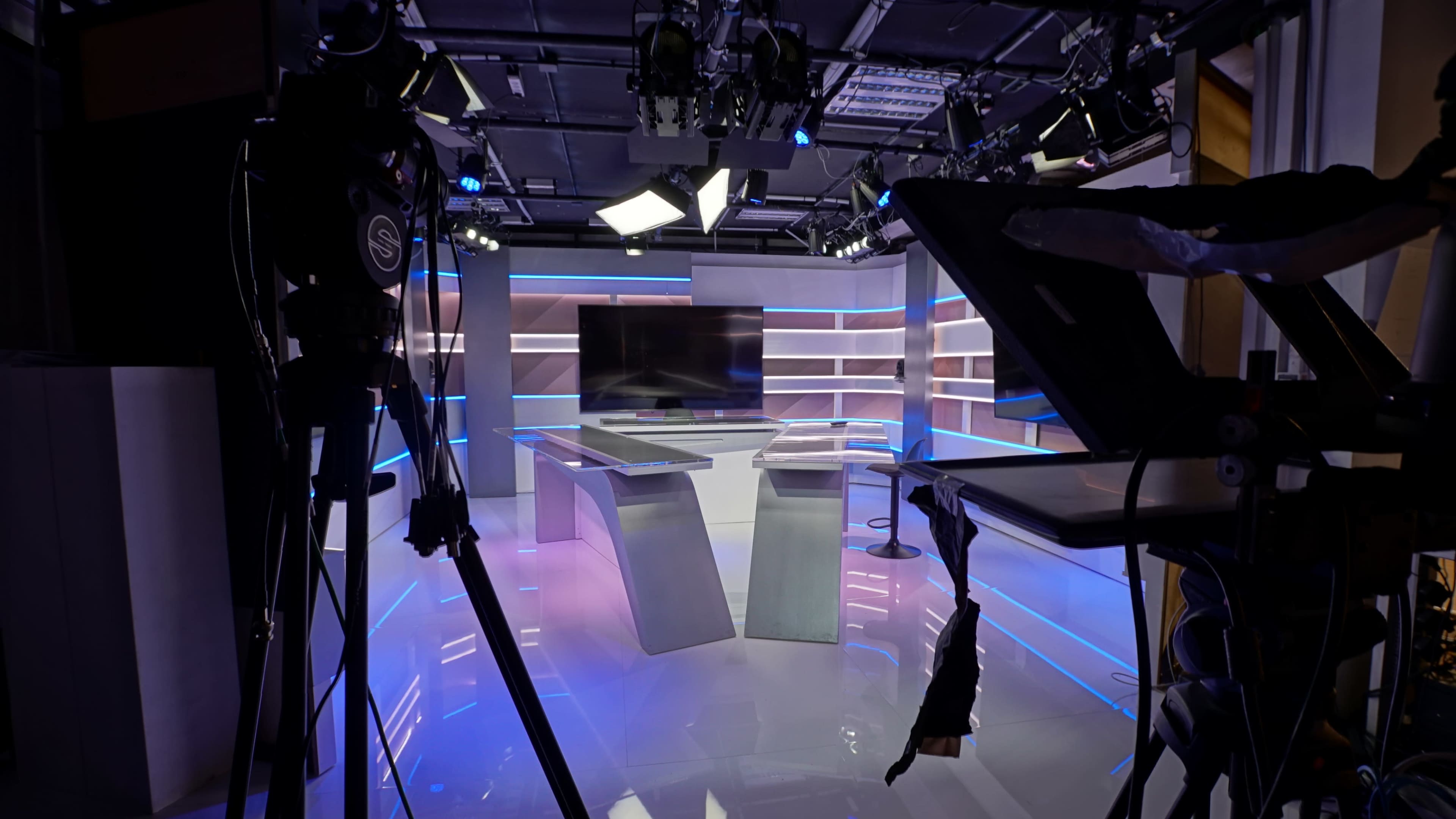 The image shows a modern empty television studio setup with a large monitor and two editing desks, surrounded by camera equipment and lighting fixtures.