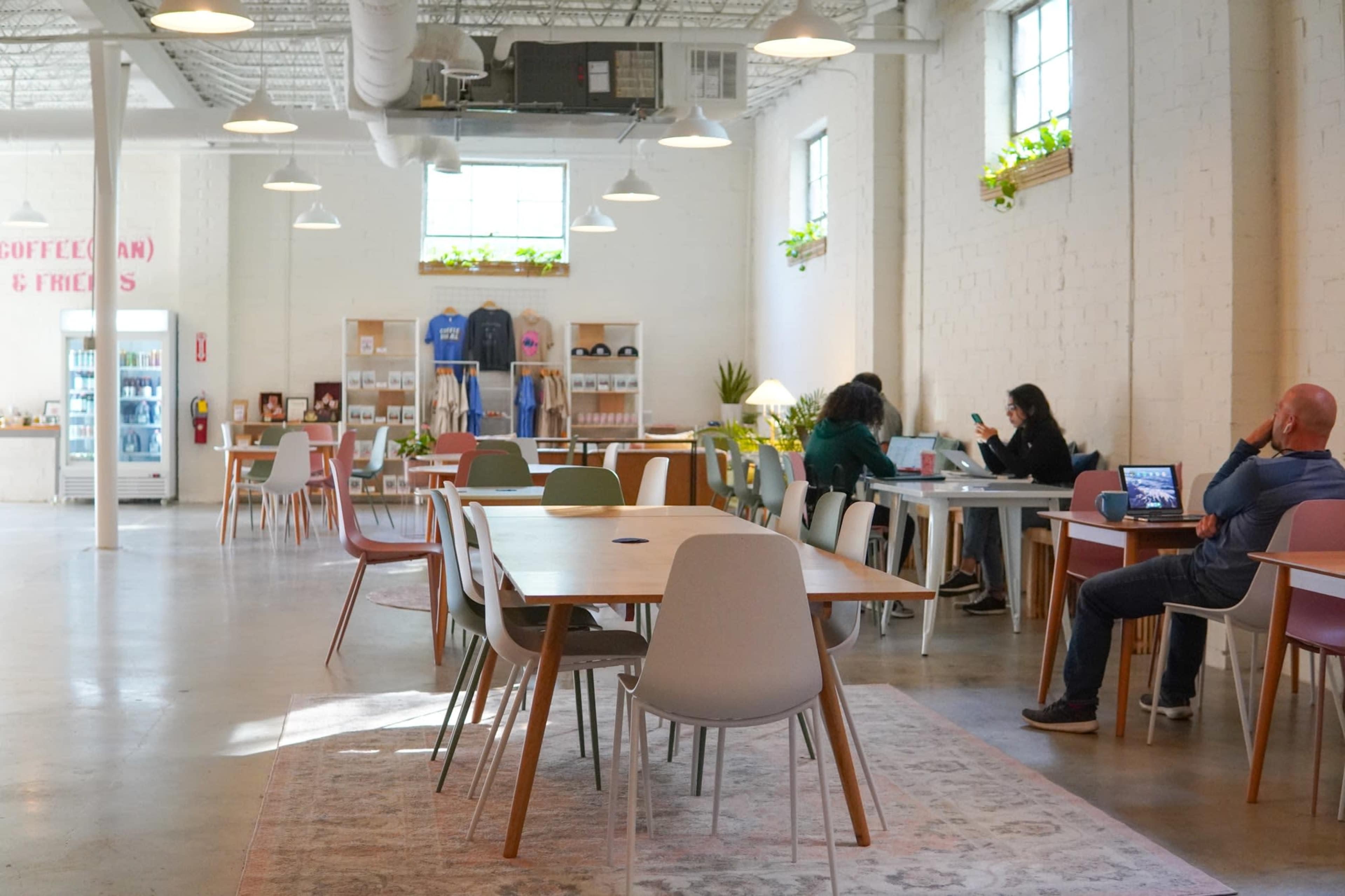 The image shows a spacious, well-lit café with wooden tables and chairs, where several people are working on laptops and others are engaged in conversation.