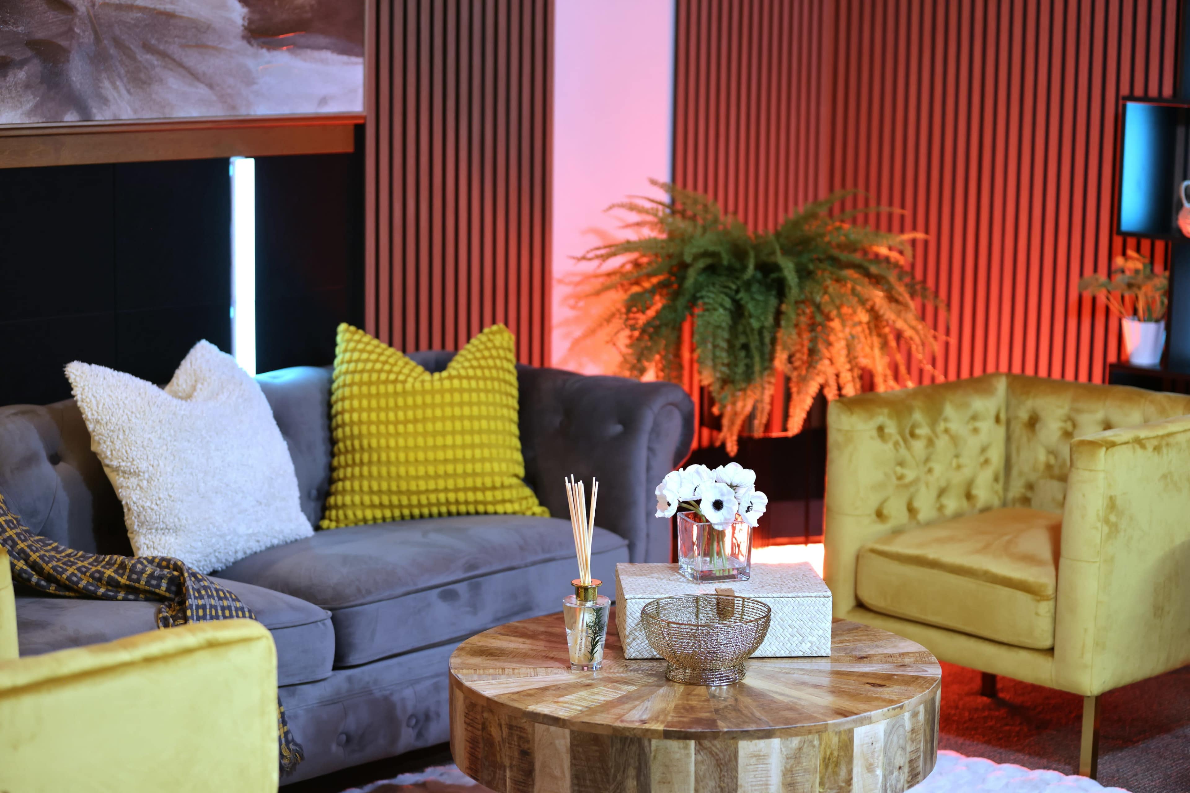 A cozy seating area features a gray sofa with colorful cushions, a wooden coffee table, and a yellow armchair, complemented by a potted fern and decorative accents.