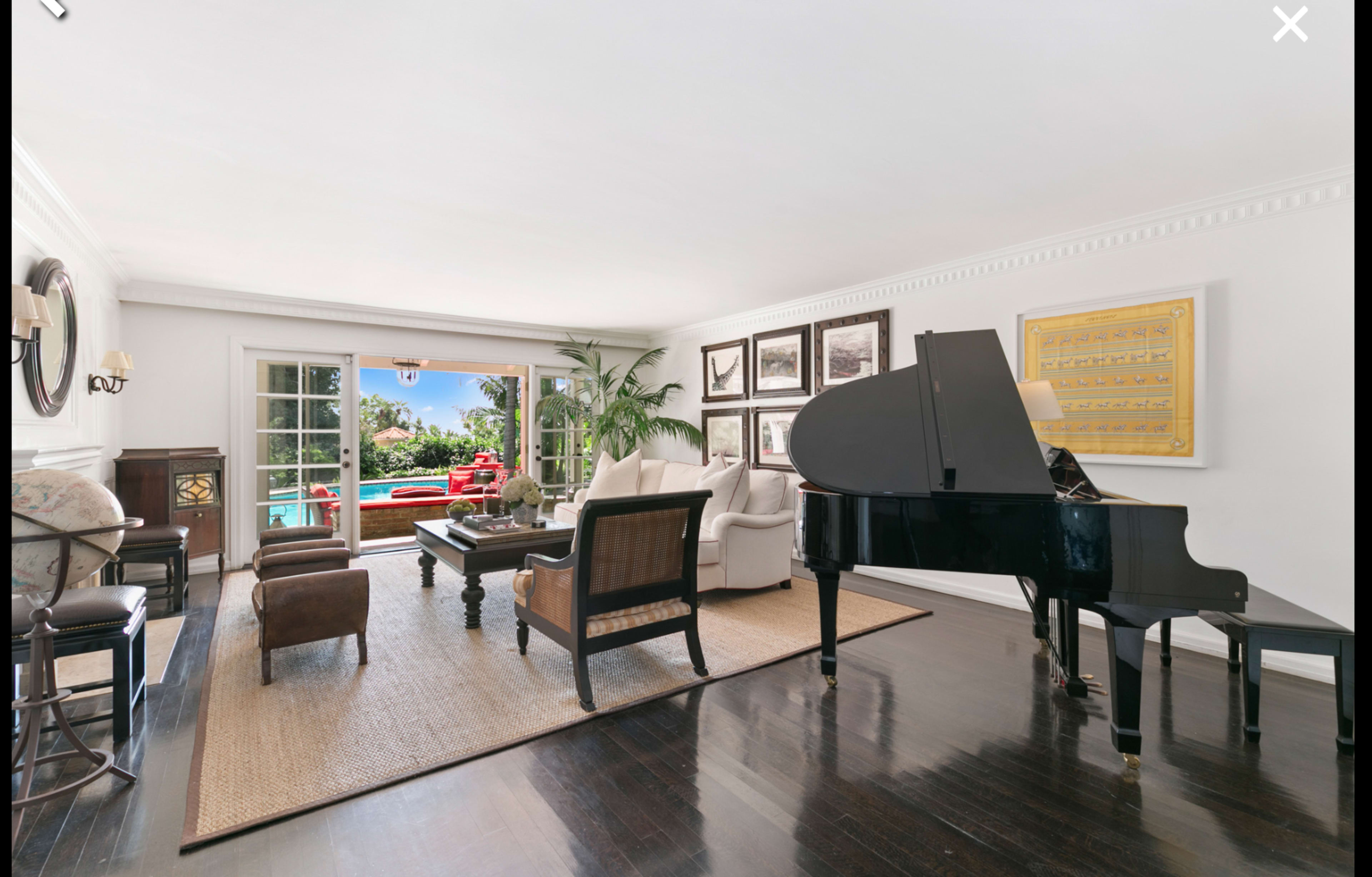 The image shows a living room featuring a grand piano, two armchairs, a coffee table, and large windows with a view of a swimming pool and greenery outside.