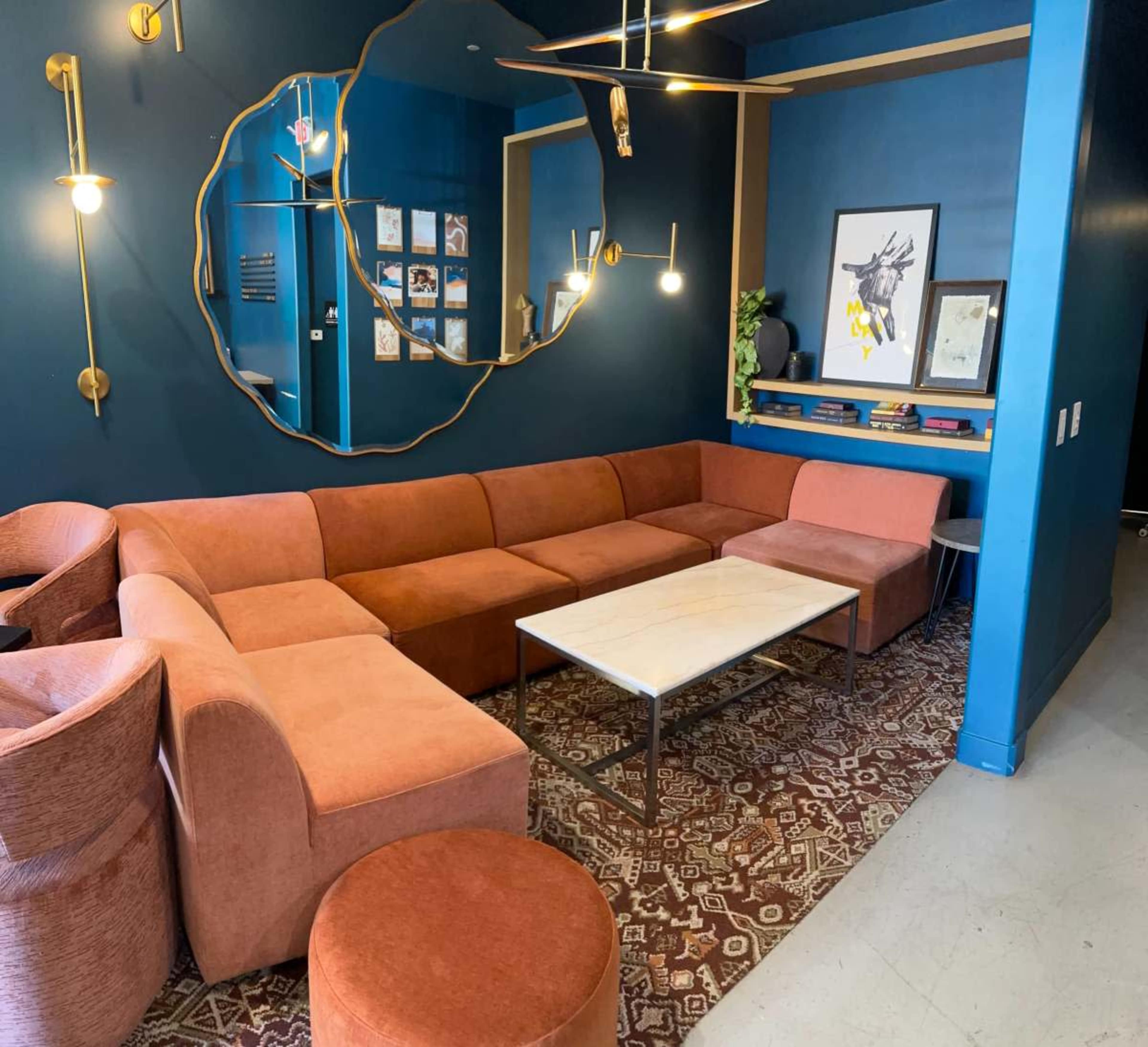 The image features a modern lounge area with a large, curved orange sectional couch, a marble coffee table, and a decorative wall mirror, set against a blue-hued wall.