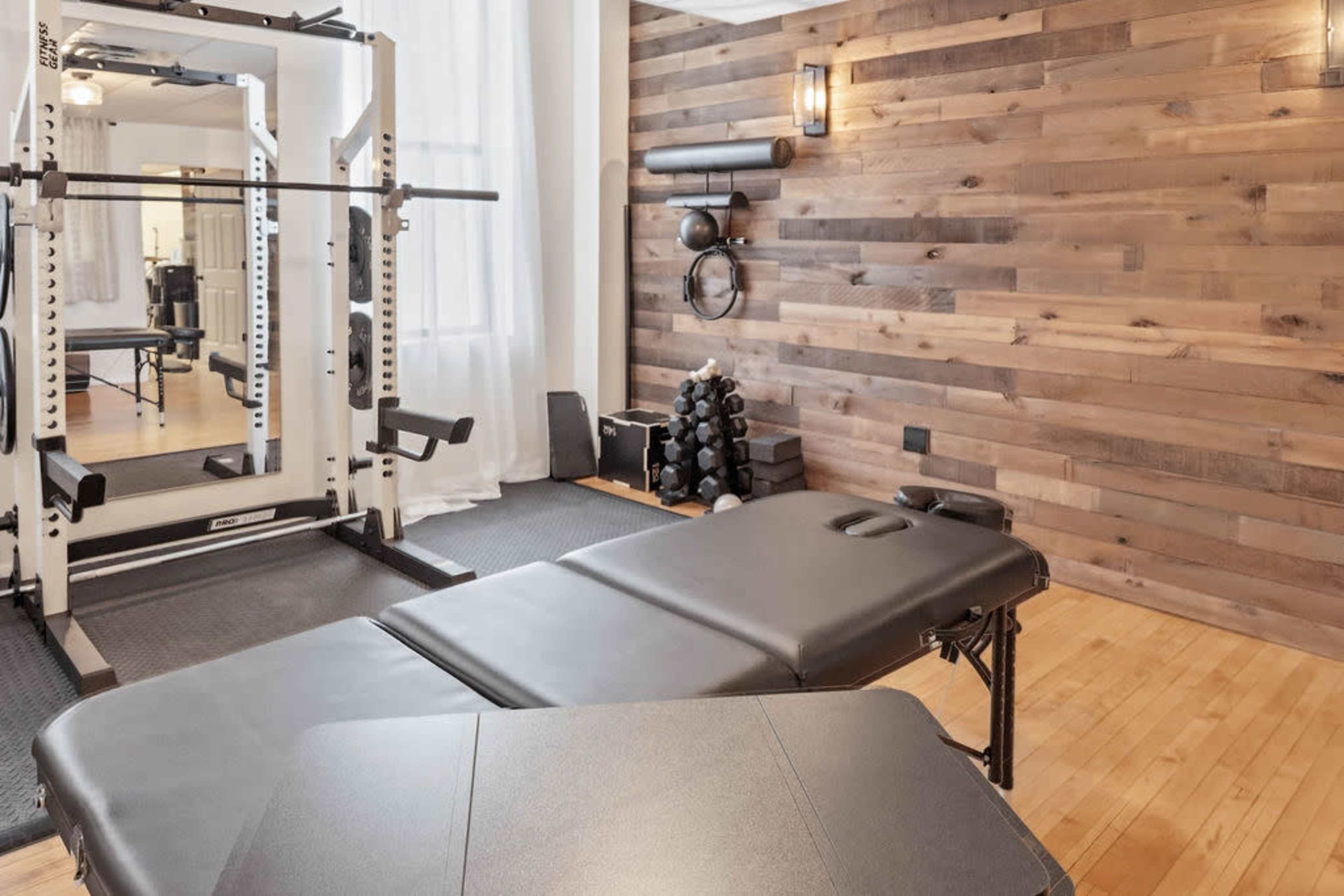 The image shows a fitness room featuring a weightlifting station, a massage table, and various exercise equipment against a wooden wall.