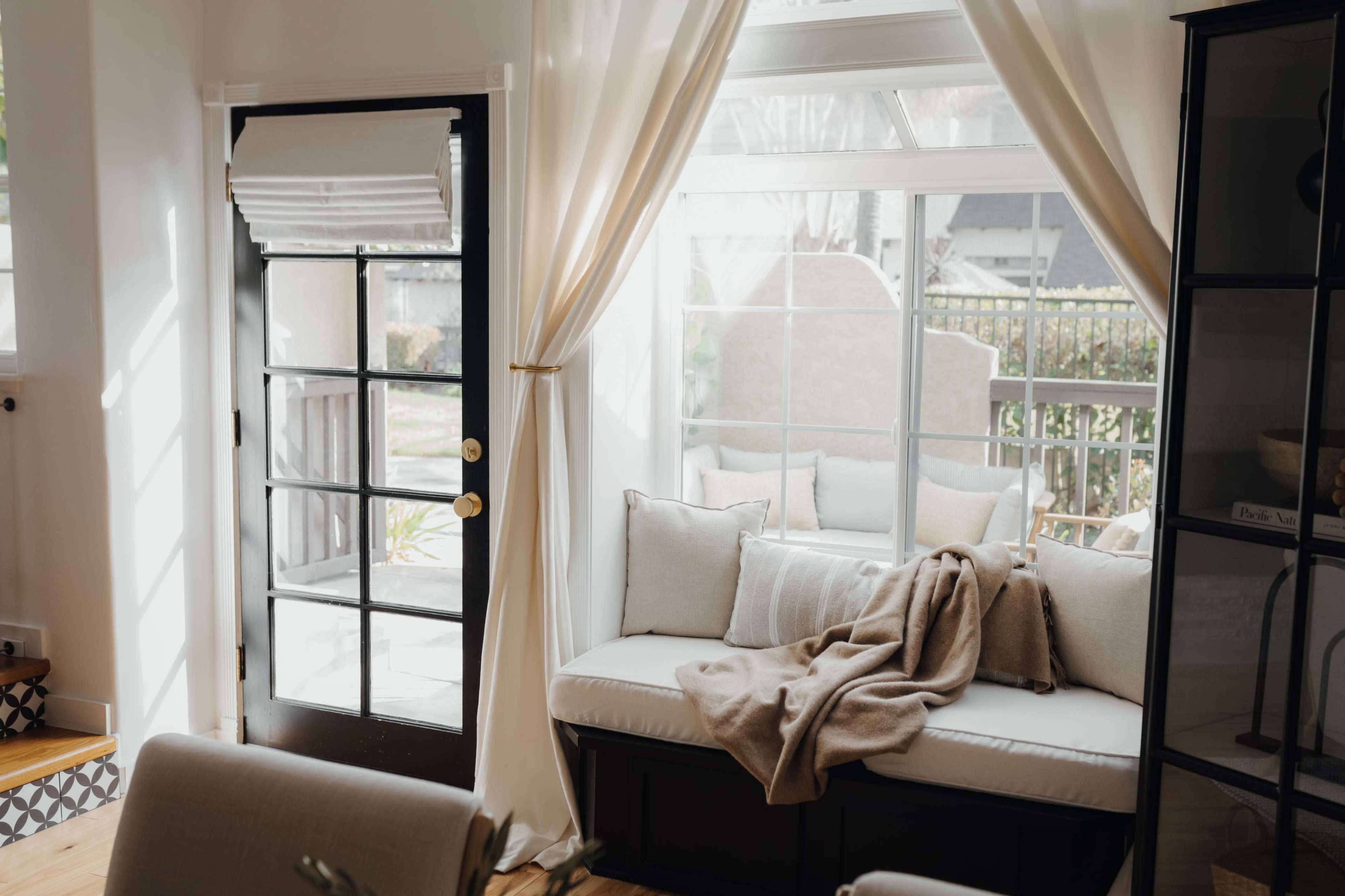 A sunlit corner of a room features a window seat with cushions and a throw blanket, framed by sheer curtains and a door leading outside.