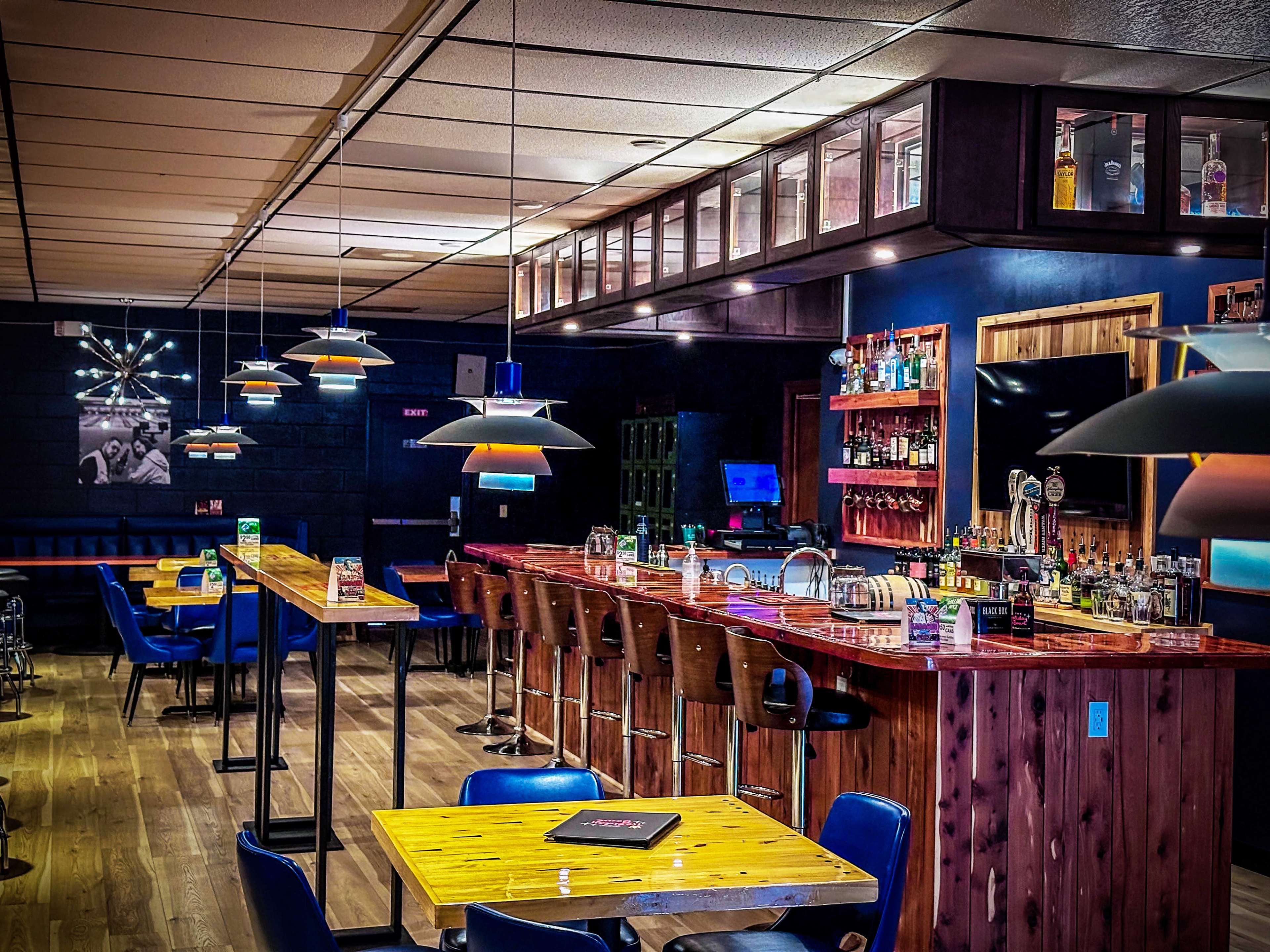 The image shows the interior of a bar with wooden tables, high chairs, and a well-stocked bar counter illuminated by pendant lights.