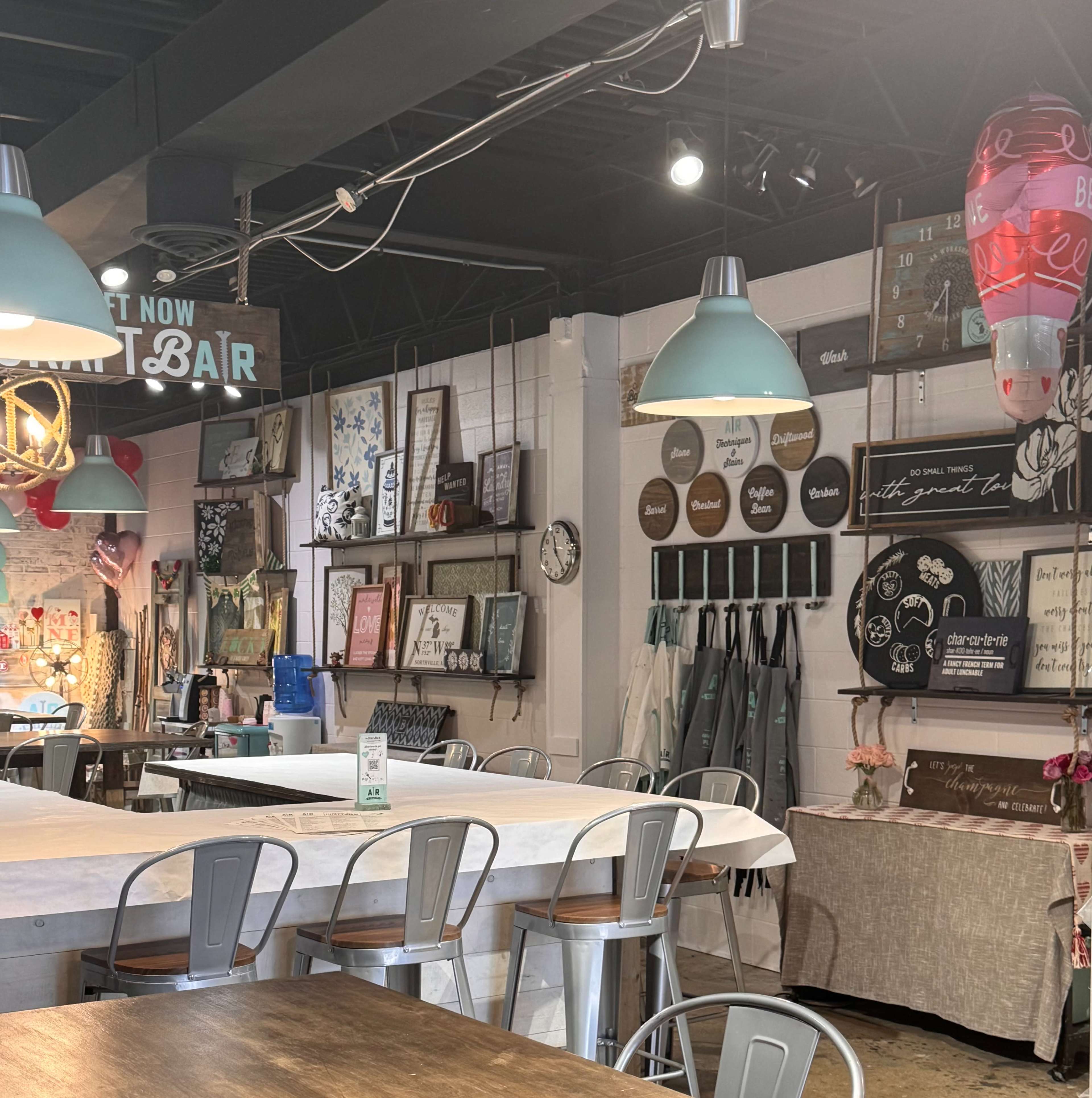 The image shows a modern craft studio with a large table, metal chairs, and various decorative signs and artwork displayed on the walls.