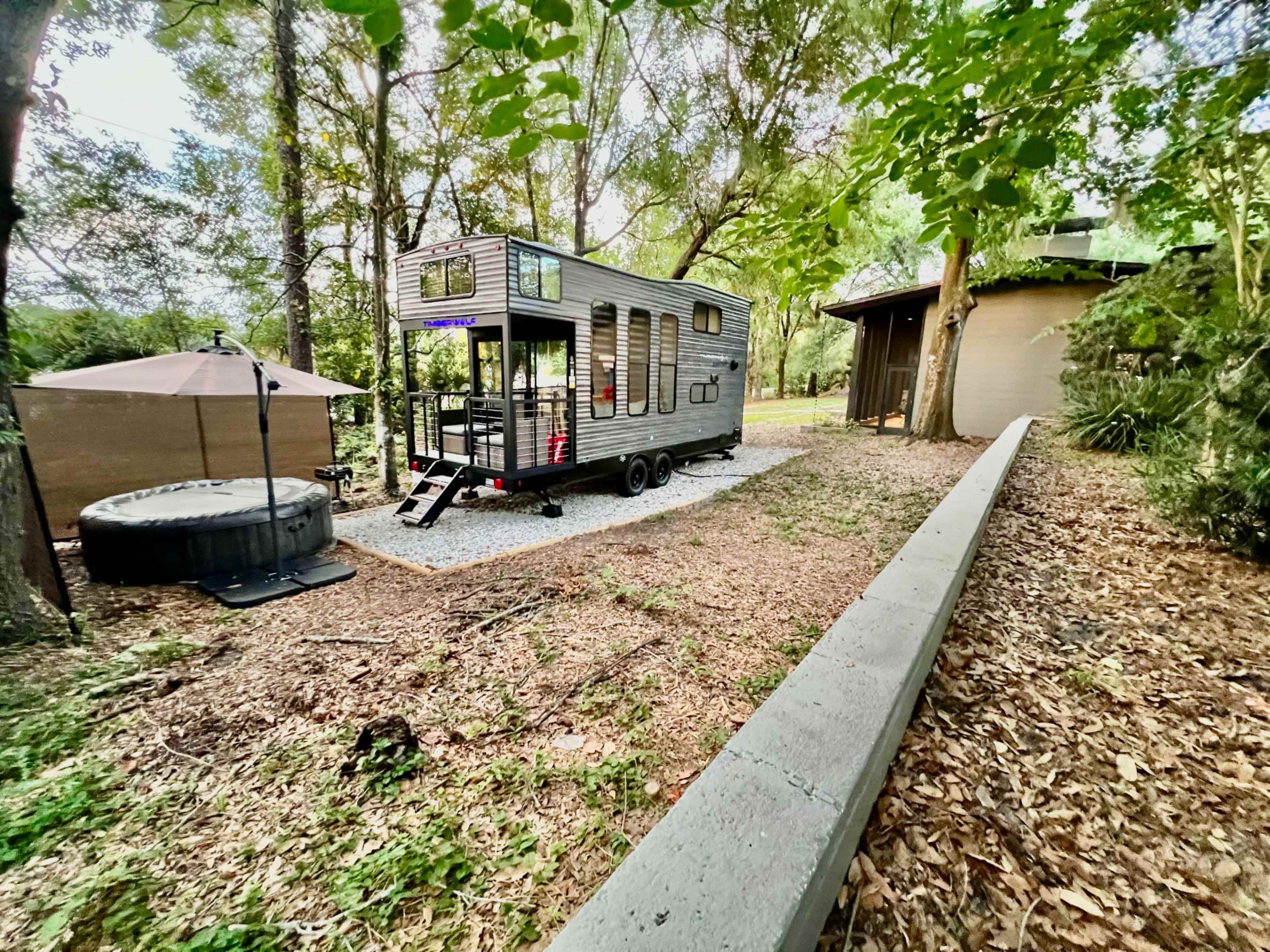 A tiny house on wheels is parked next to a circular building, surrounded by trees and a gravel path.
