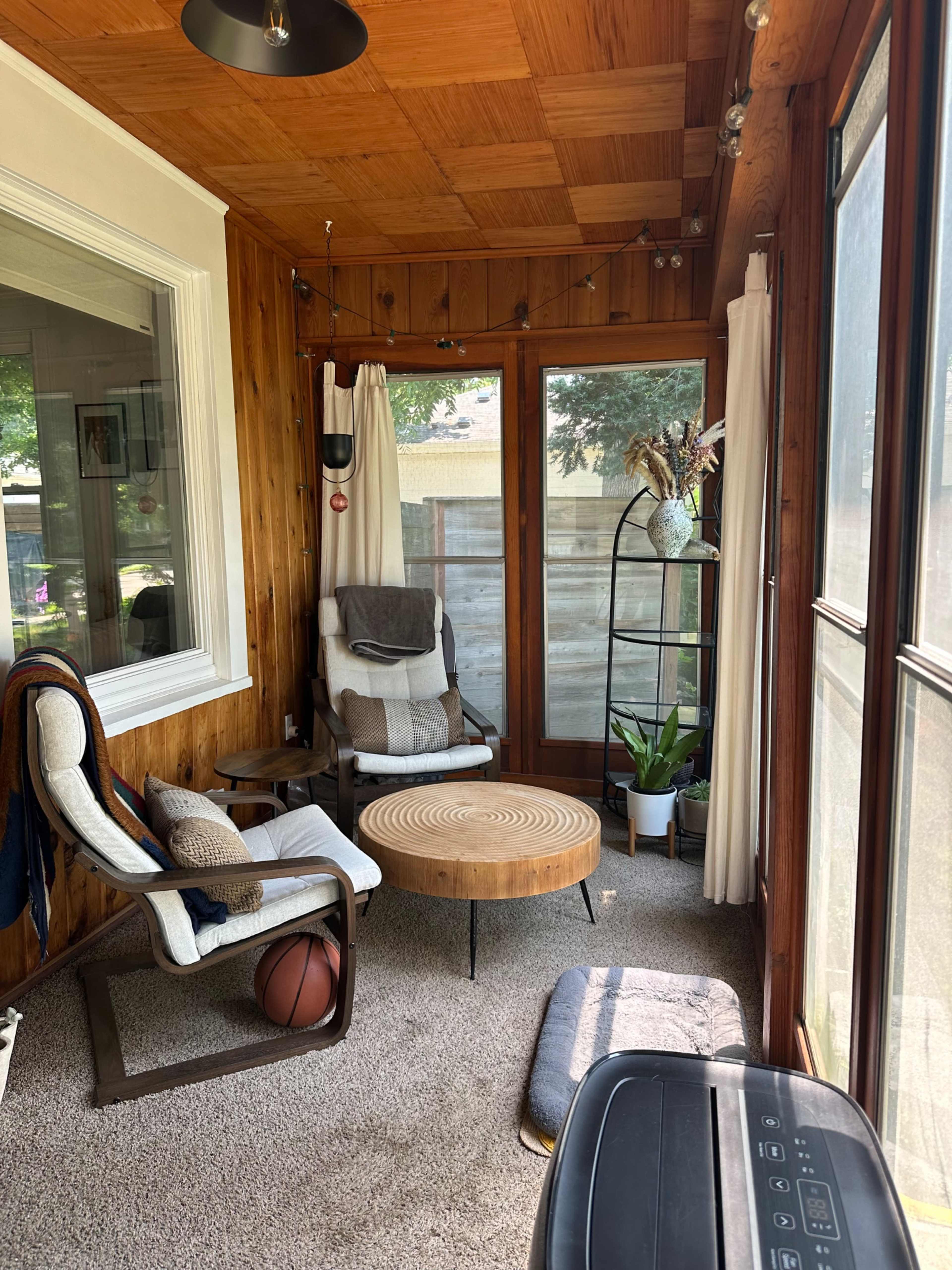 The image shows a cozy sunroom with wooden walls, a circular wooden coffee table, two lounge chairs, a decorative plant, and large windows letting in natural light.