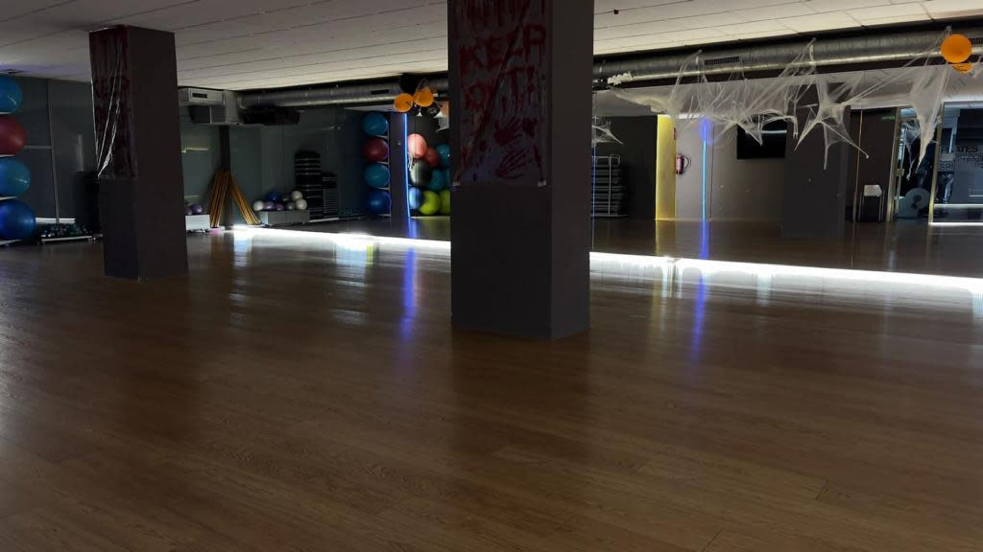 The image shows an empty gym studio with wooden flooring, mirrored walls, and exercise equipment along the sides, illuminated by lights.