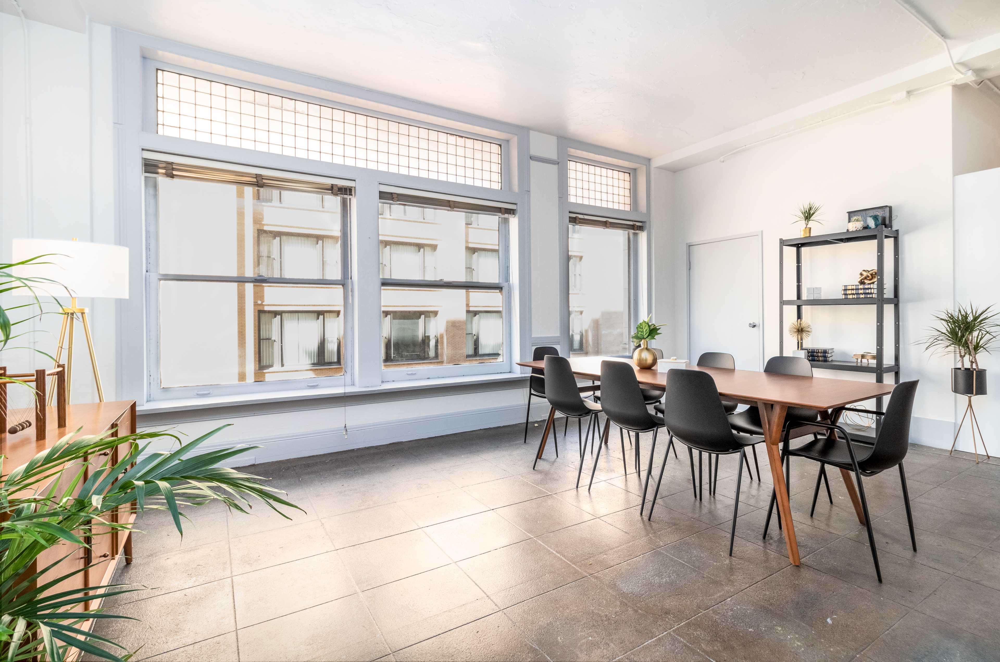 The image shows a bright meeting room with large windows, a long table surrounded by black chairs, and a shelving unit in the corner.